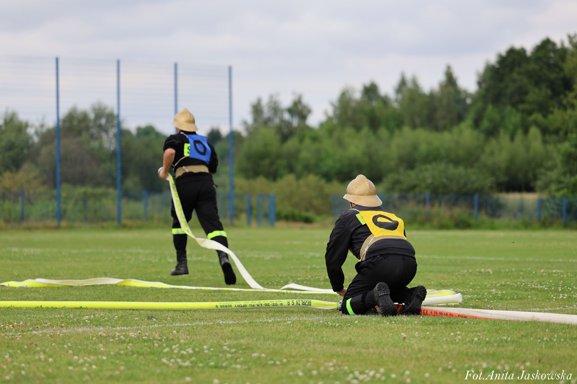 Dwóch strażaków w hełmach i czarnych uniformach z kolorowymi oznaczeniami rozkłada wąż strażacki na zielonym boisku z siatką w tle.