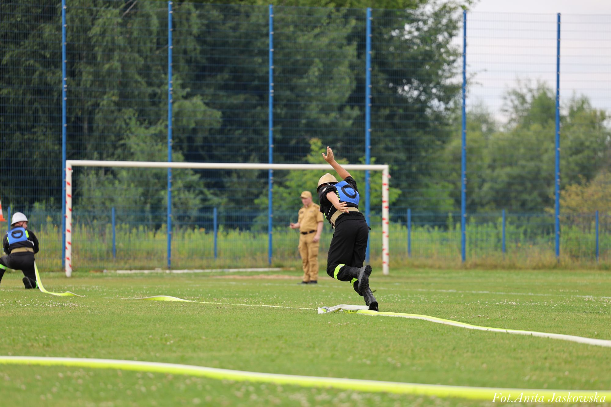 Dwie osoby w czarnych strojach z niebieskimi numerami i hełmach na zielonym boisku, jedna biegnie, druga kuca, w tle sędzia w beżowym ubraniu i bramka piłkarska.