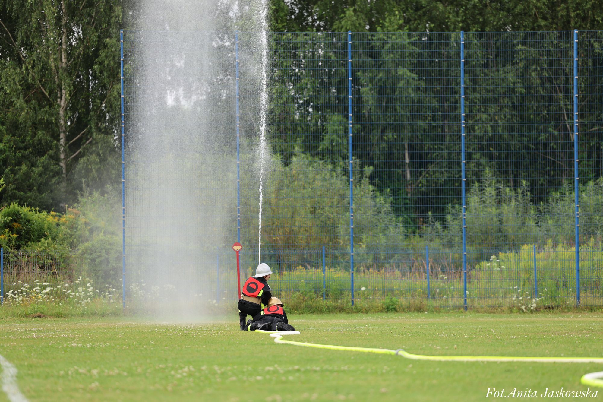 Dwoje osób w hełmach strażackich i czerwonych kamizelkach operuje strumieniem wody z węża na zielonym boisku, w tle niebieski ogrodzenie i drzewa.