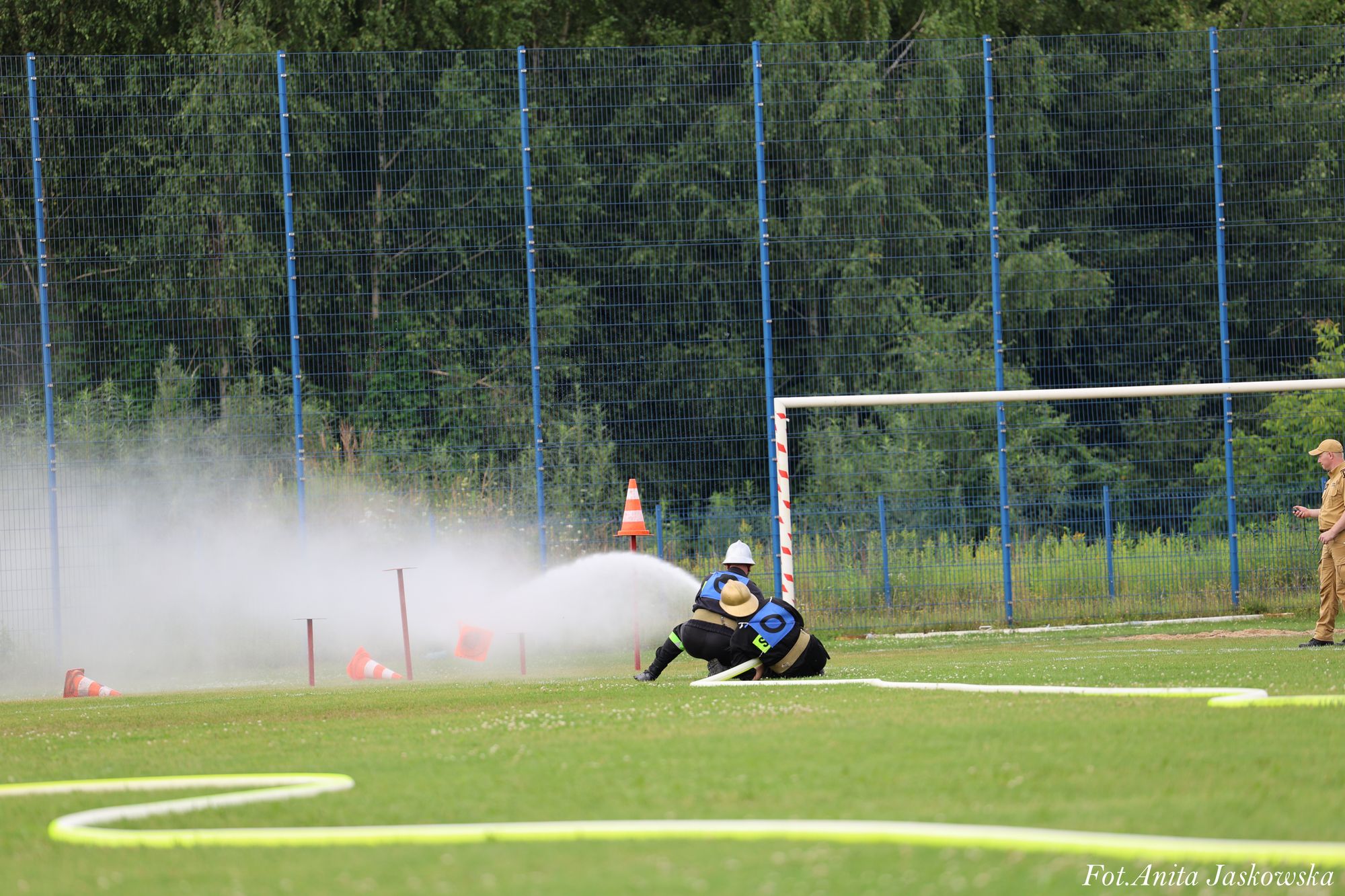 Dwie osoby w ubraniach ochronnych z niebieskimi oznaczeniami obsługują strumień wody z węża strażackiego na zielonym boisku sportowym, w tle niebieski ogrodzenie i drzewa.