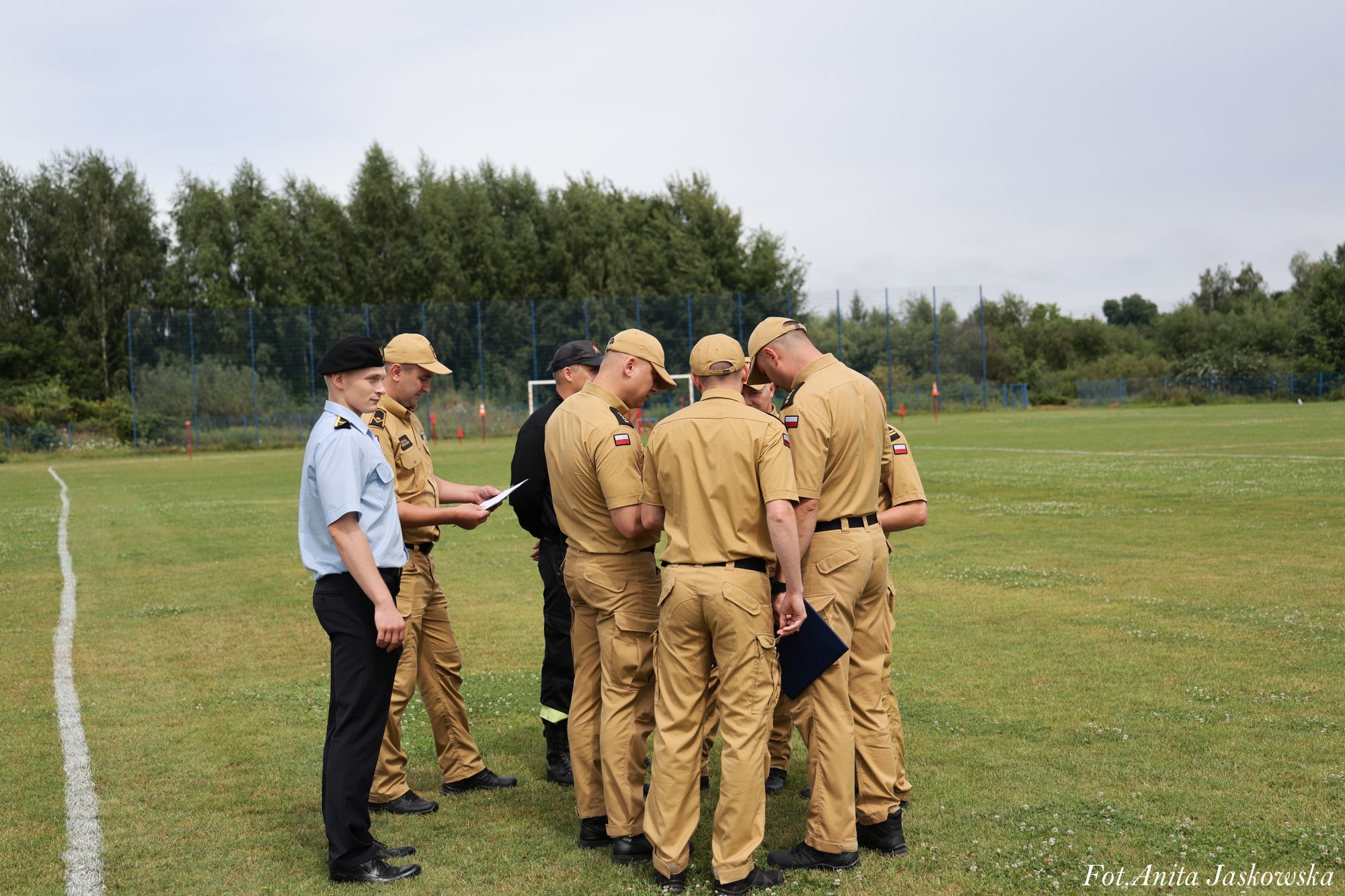 Grupa mężczyzn w mundurach beżowych i niebieskim stojąca na zielonej trawie na boisku, w tle drzewa i niebieskie ogrodzenie.