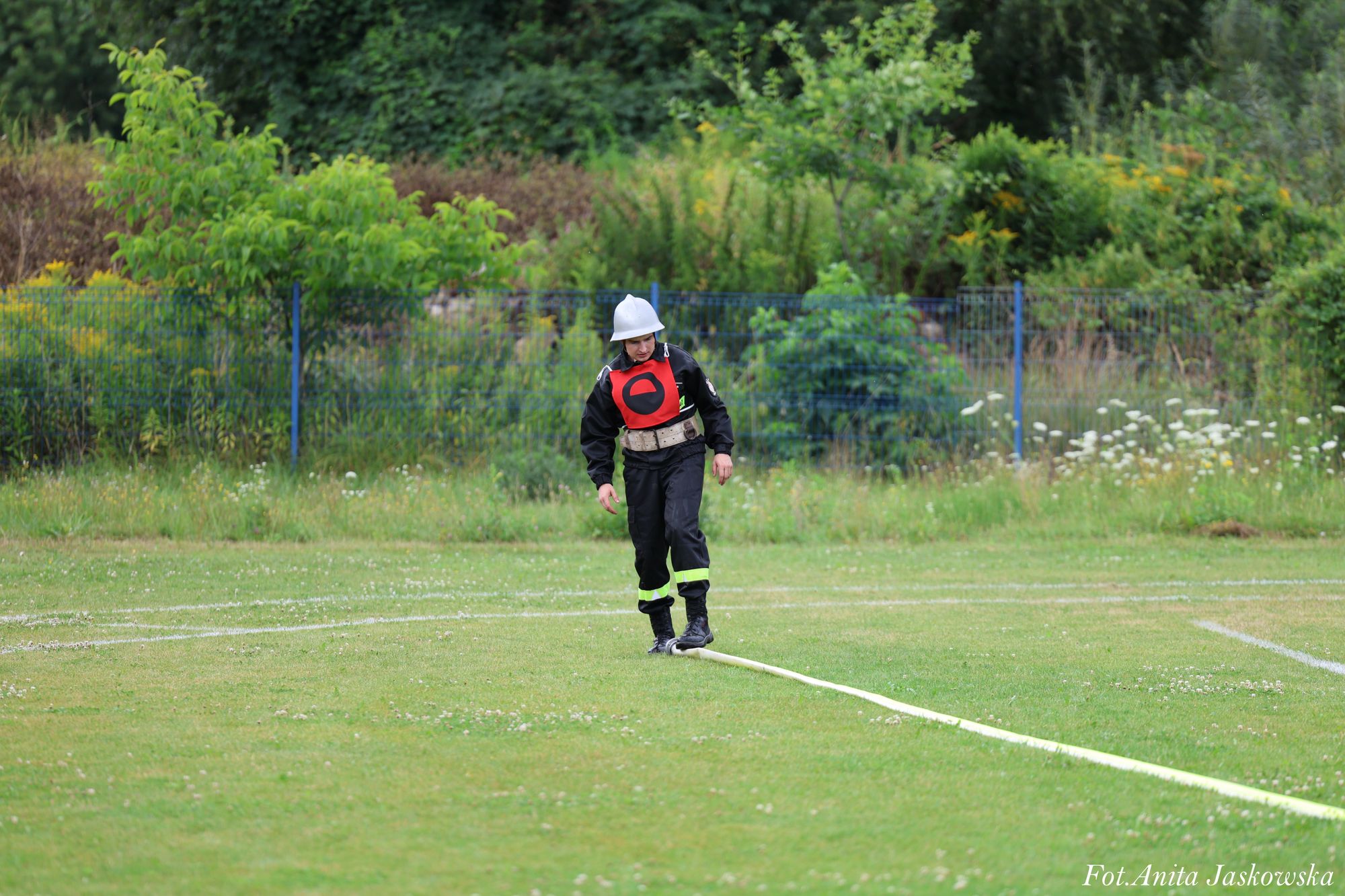Strażak w czarnym ubraniu i białym kasku stoi na zielonej trawie, obok rozciągnięty jasny wąż strażacki, w tle zielone krzewy i metalowy płot.