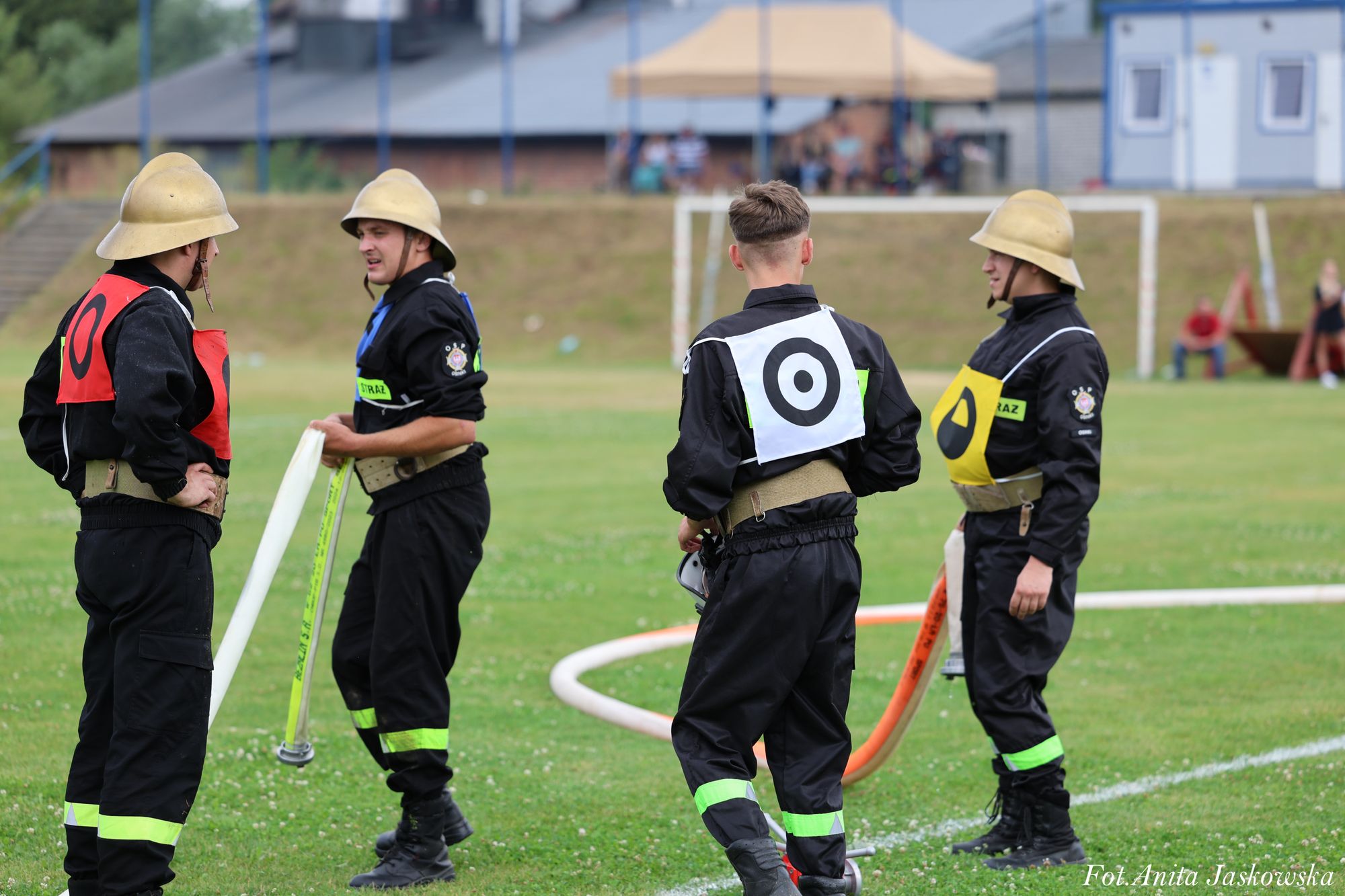 Czterech młodych strażaków w czarnych mundurach z odblaskowymi paskami i hełmami stojących na trawie, trzymających węże strażackie, w tle boisko i budynki.