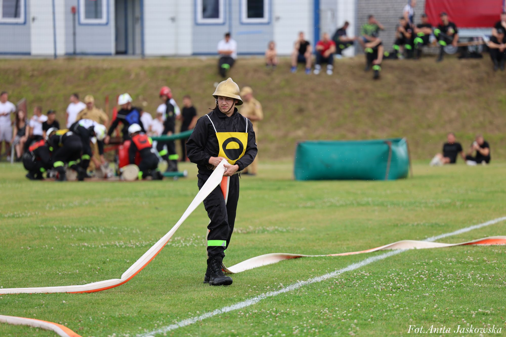 Strażak w czarnym ubraniu i hełmie przeciąga biało-pomarańczowy wąż na zielonym boisku, w tle grupa osób i budynki.