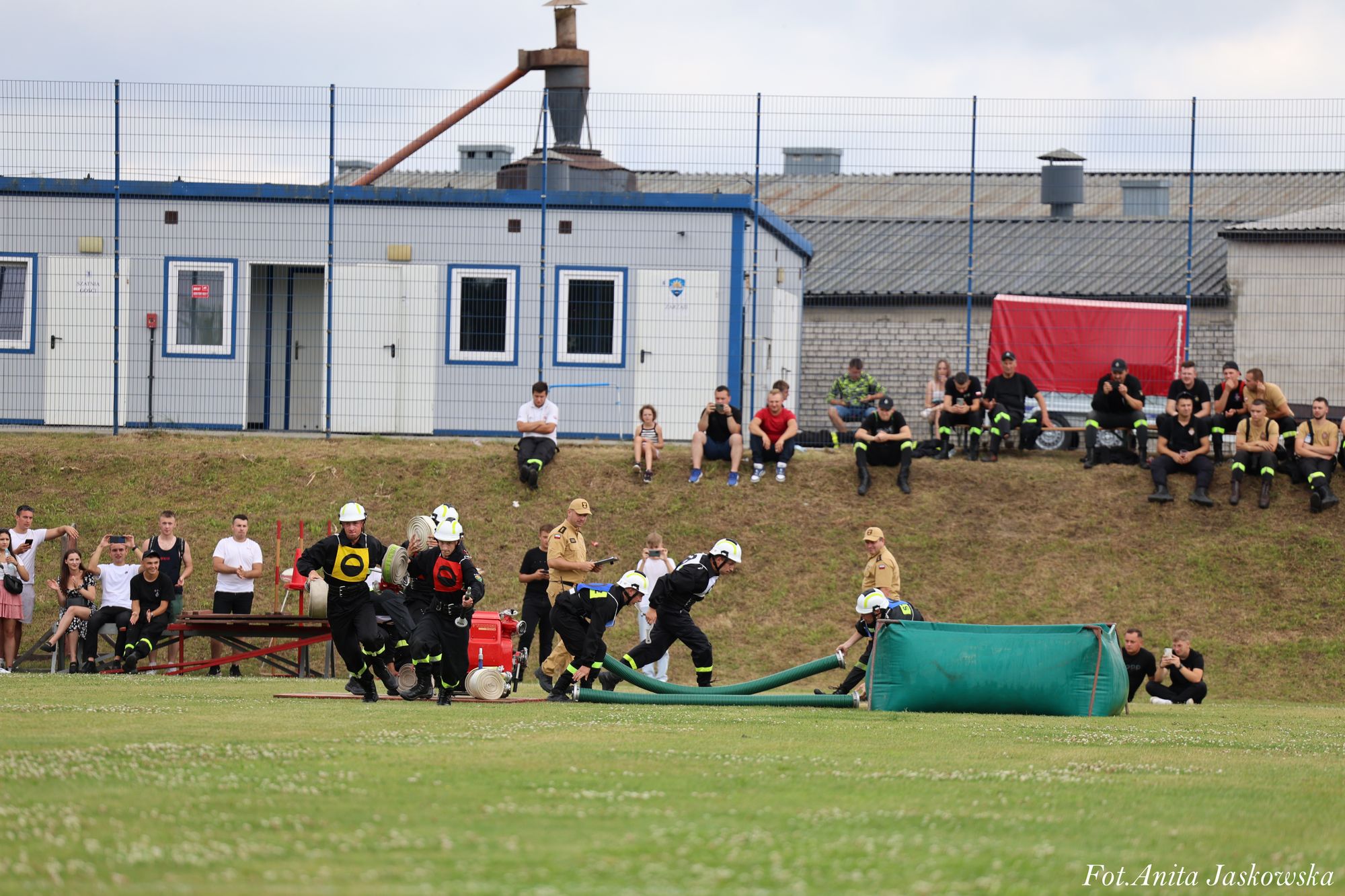 Grupa strażaków w czarnych mundurach i białych hełmach podczas ćwiczeń na zielonym boisku, w tle ludzie siedzący na trawie i budynki za ogrodzeniem.
