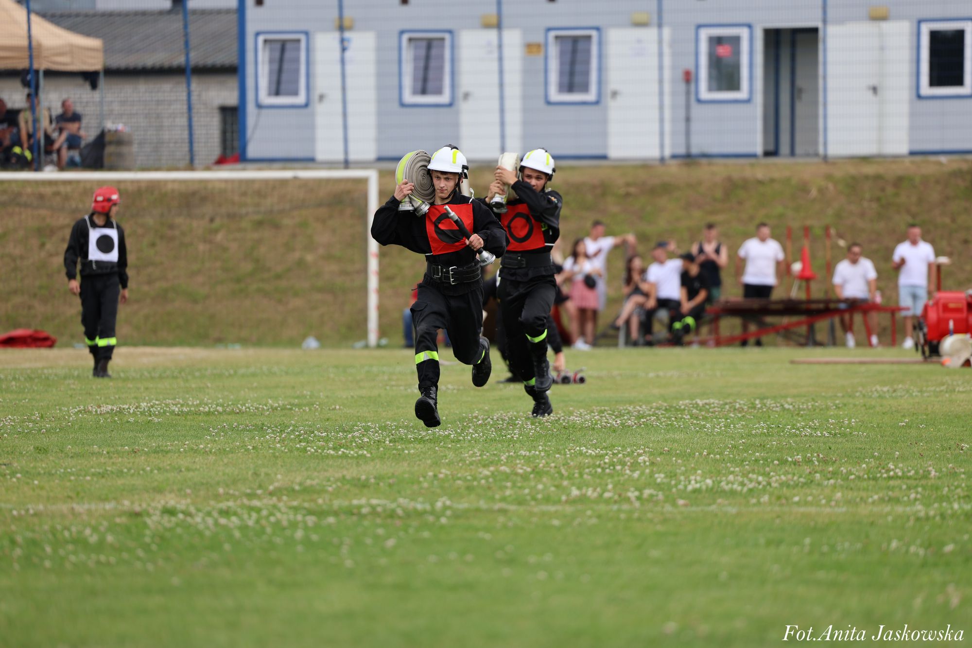 Dwóch strażaków w kaskach i czarnych mundurach z czerwonymi oznaczeniami biegnie po zielonym boisku, każdy niesie zwinięty wąż strażacki, w tle ludzie i budynki.
