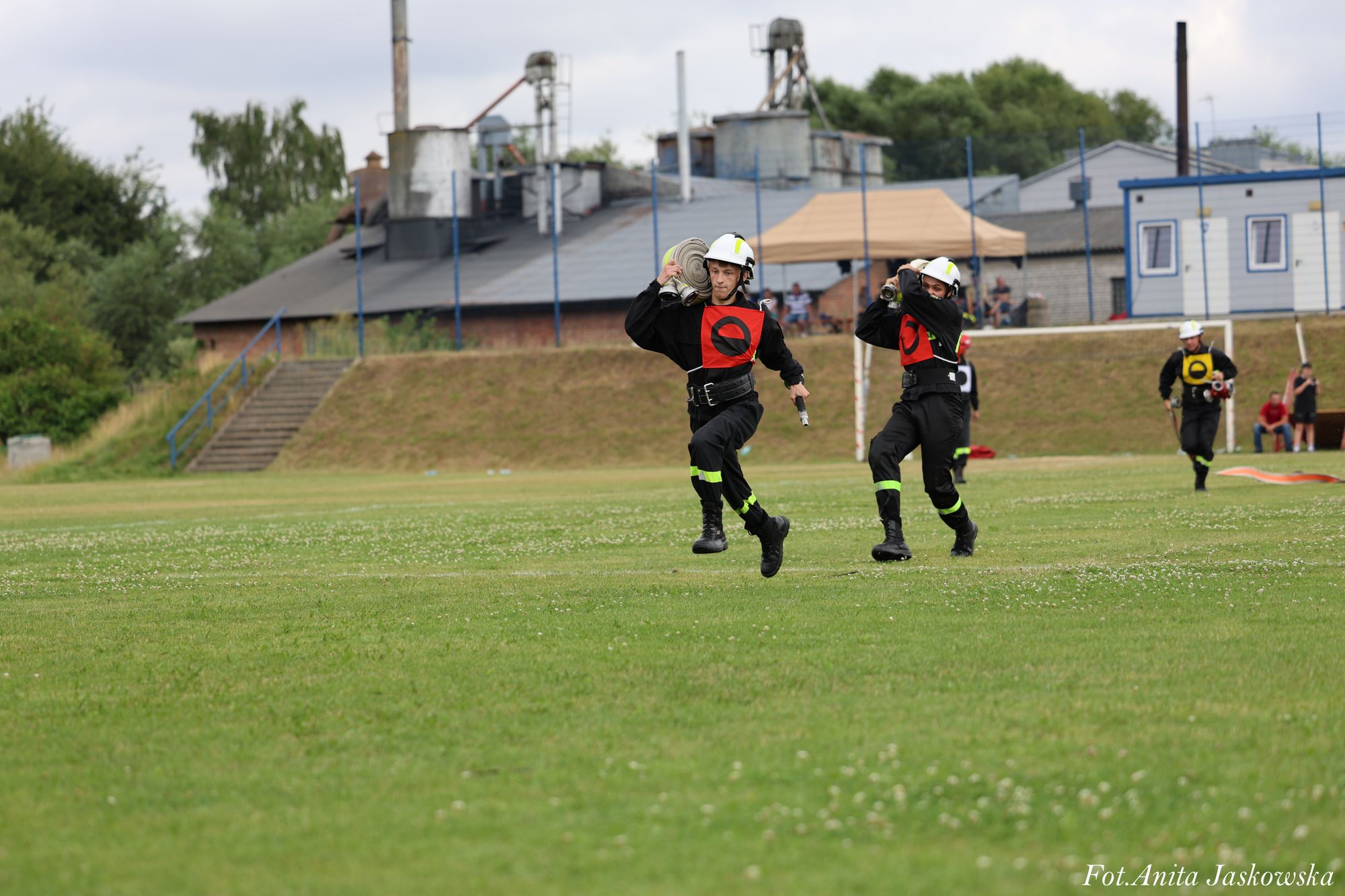 Trzech strażaków w hełmach i czarnych uniformach z czerwonymi i żółtymi oznaczeniami biegnie po zielonym trawiastym boisku, niosąc zwinięte węże strażackie.