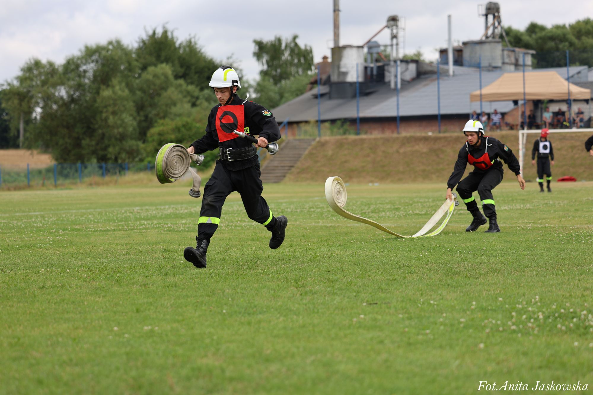 Dwóch strażaków w czarnych mundurach i białych hełmach z czerwonymi numerami, biegnących po zielonym boisku, trzymających rozwinięte węże strażackie.