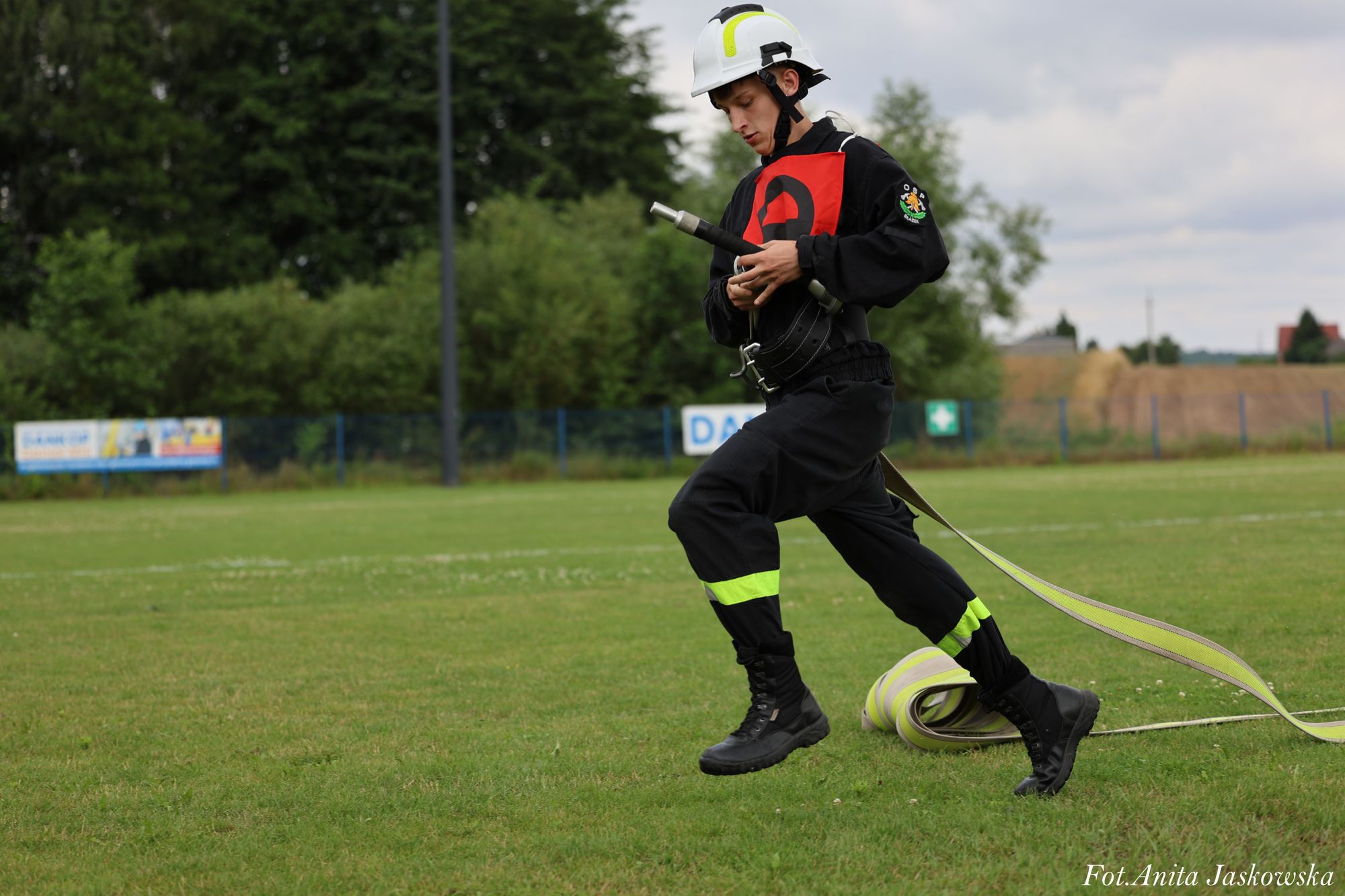 Młody strażak w czarnym mundurze z odblaskowymi elementami i białym hełmem biegnie po zielonym boisku trzymając zwinięty wąż strażacki.