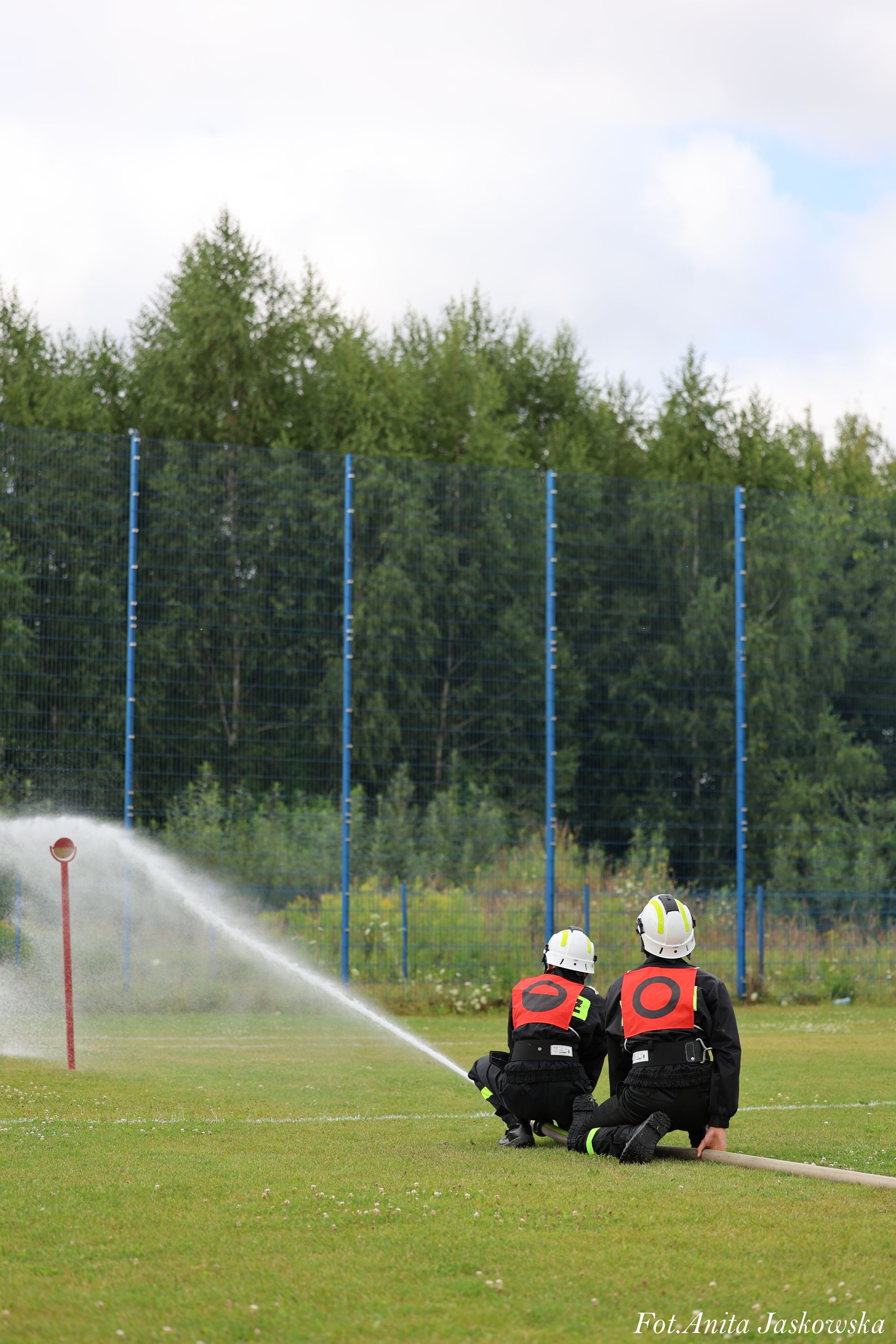 Dwóch strażaków w czarnych mundurach i białych hełmach z czerwonymi oznaczeniami, klęczących na zielonej trawie, trzymających wąż gaśniczy z wystrzeliwującą wodą, na tle ogrodzenia i drzew.