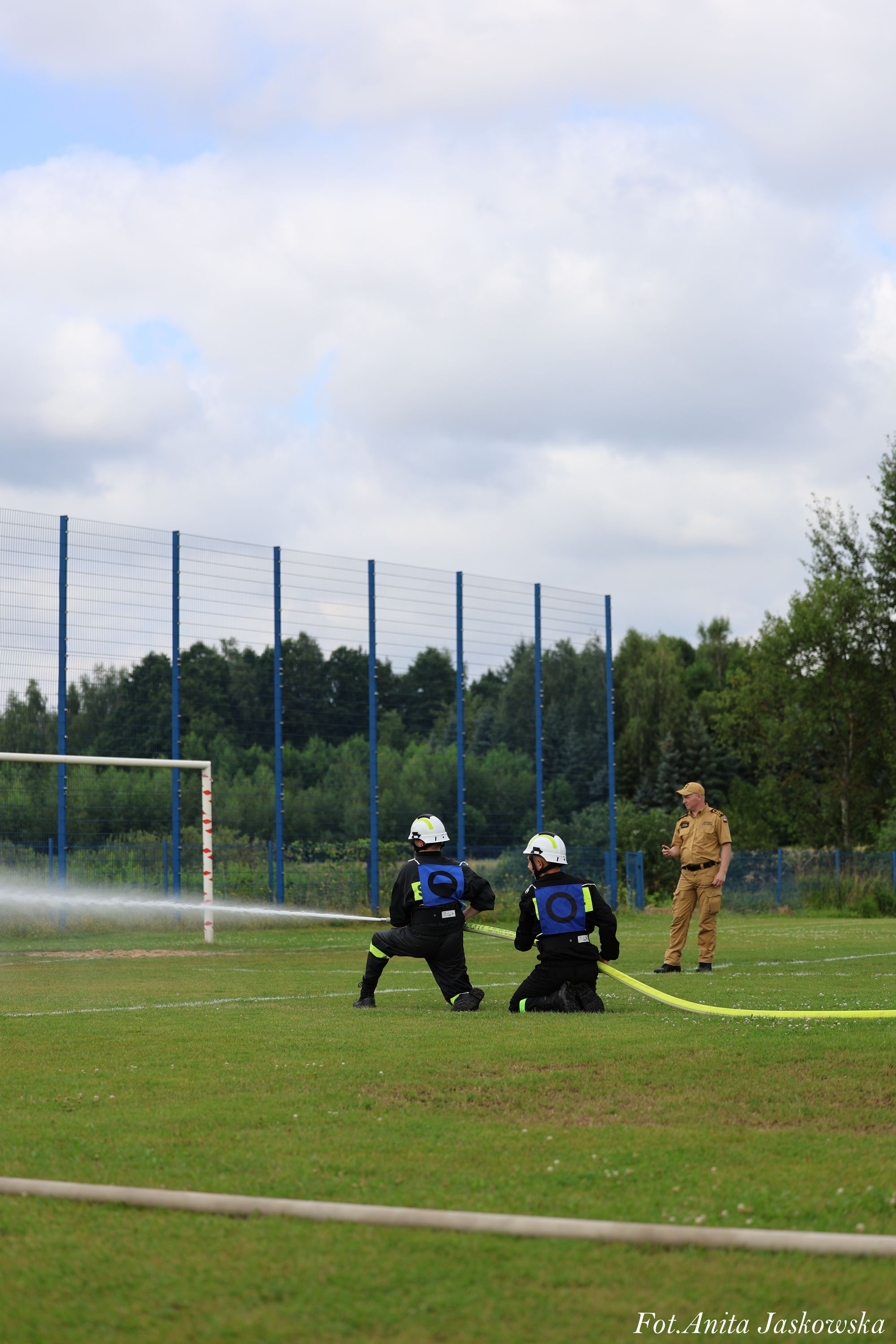 Dwóch strażaków w czarnych mundurach i białych hełmach z wężem gaśniczym na zielonym boisku sportowym, obok stoi mężczyzna w brązowym mundurze.