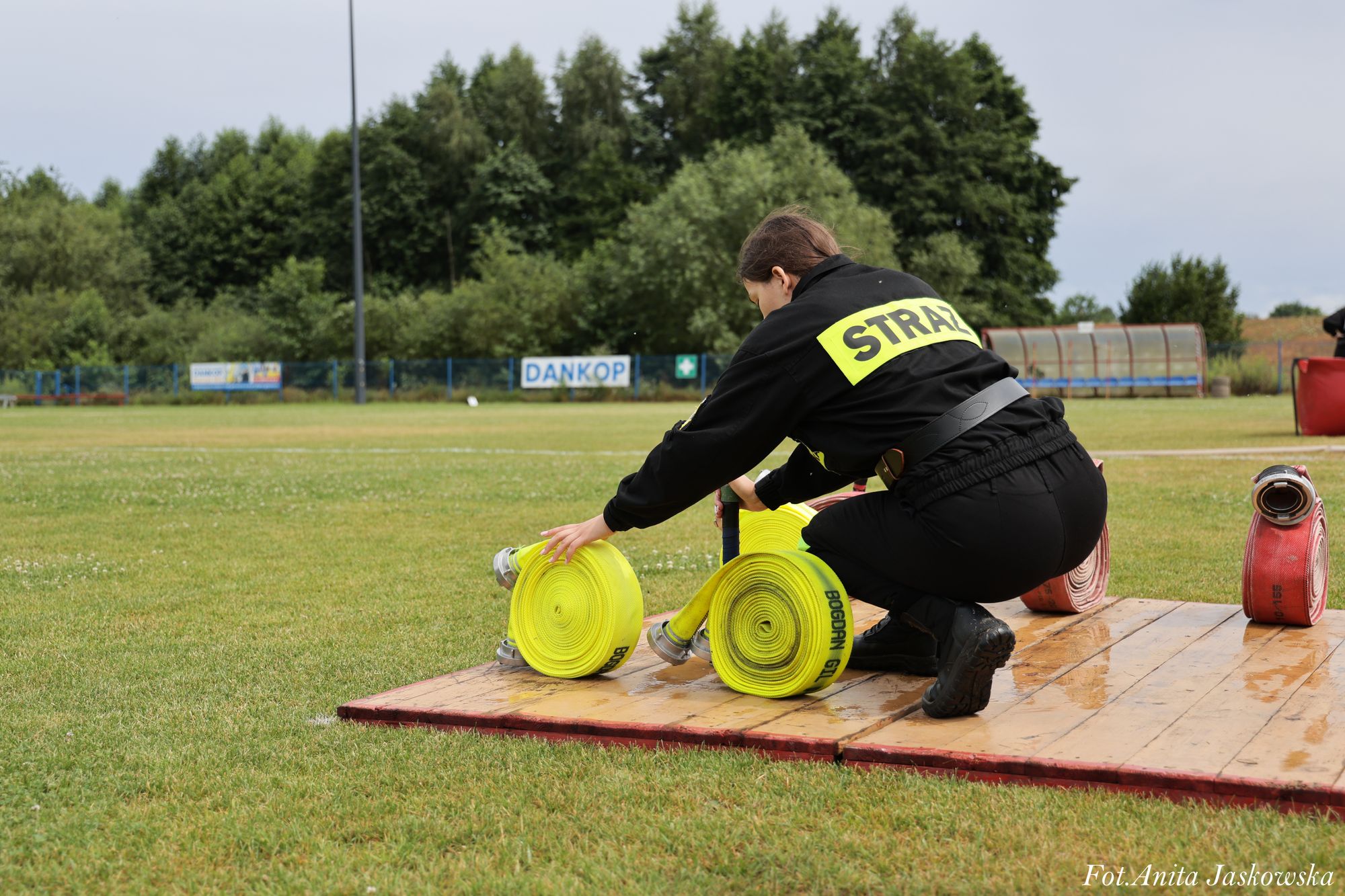 Strażak w czarnym ubraniu z napisem STRAŻ zwija żółte węże strażackie na drewnianej platformie na zielonym boisku z drzewami w tle.