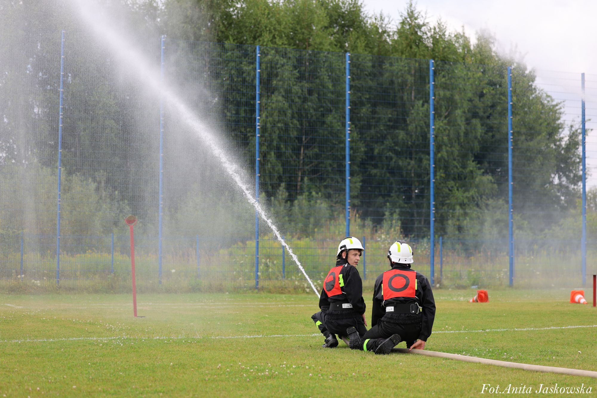 Dwóch strażaków w czarnych uniformach i białych hełmach klęczy na zielonej trawie, jedna z nich trzyma wąż strażacki z którego leci strumień wody.