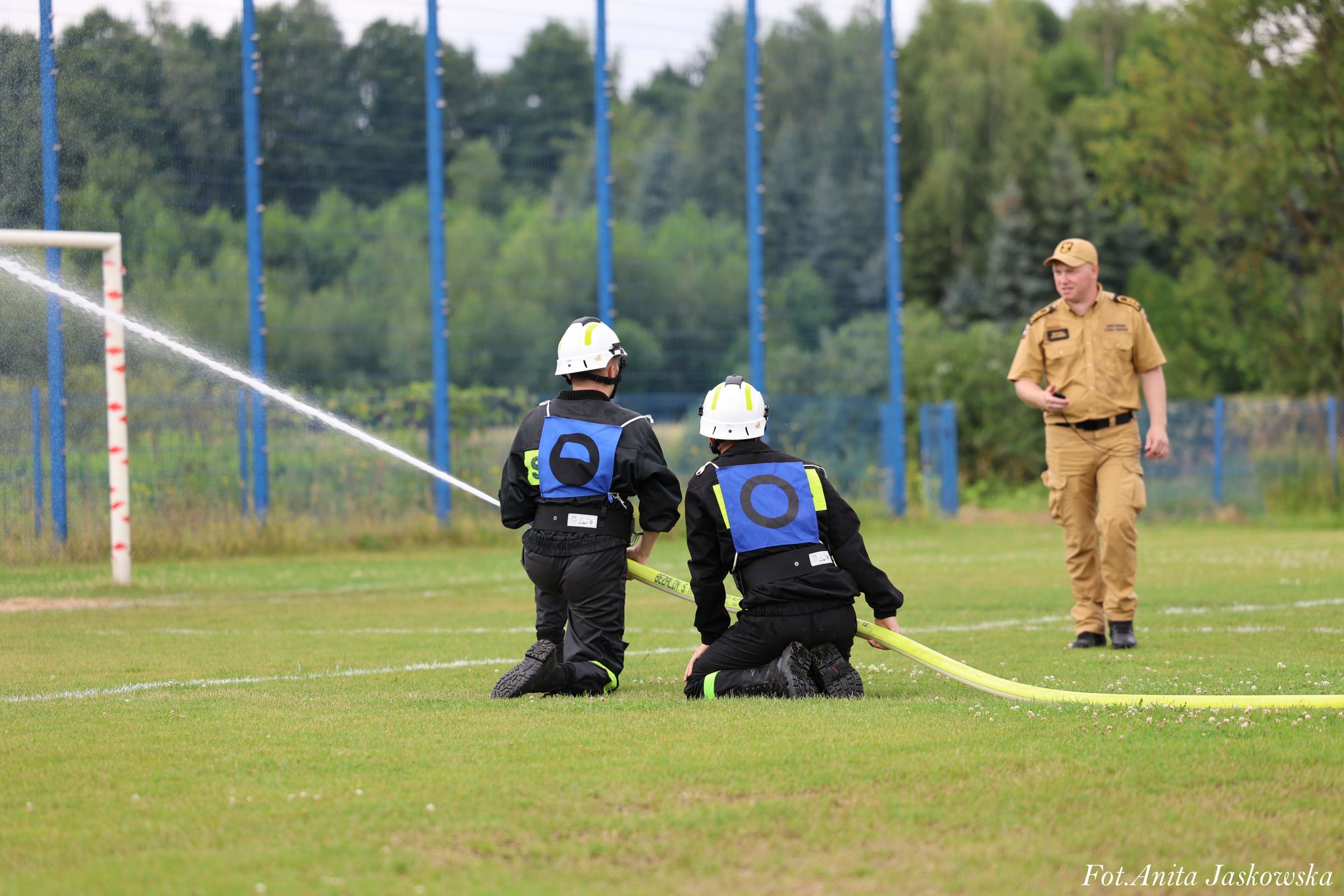 Dwóch strażaków w kaskach i czarnych mundurach z niebieskimi oznaczeniami klęczy na zielonym boisku, trzymając żółty wąż strażacki z wypływającą wodą, obok stoi mężczyzna w beżowym mundurze.