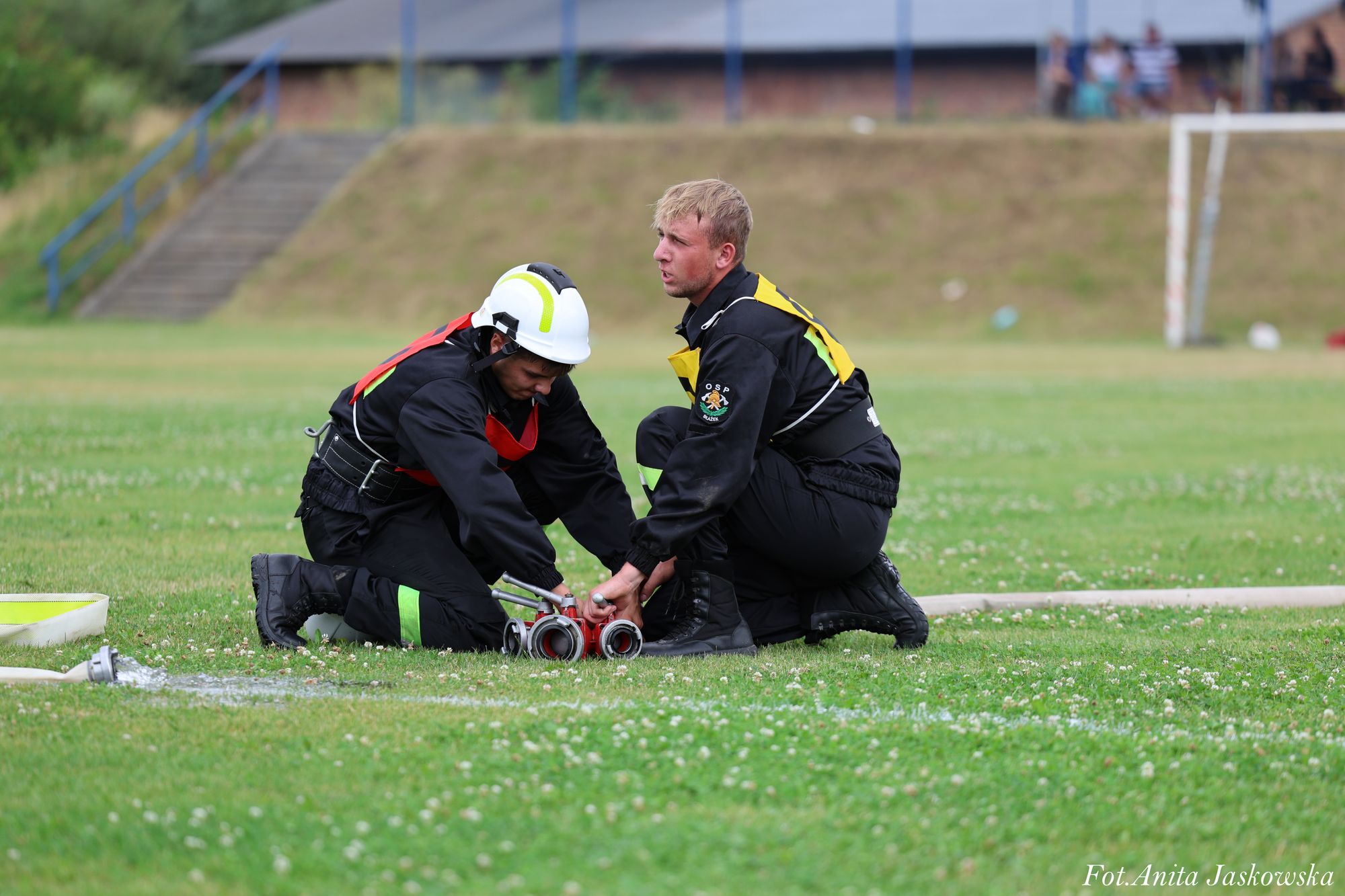 Dwóch mężczyzn w czarnych mundurach strażackich z kolorowymi oznaczeniami, jeden w białym hełmie, klęczy na zielonej trawie, obsługując czerwony sprzęt z wężami strażackimi.