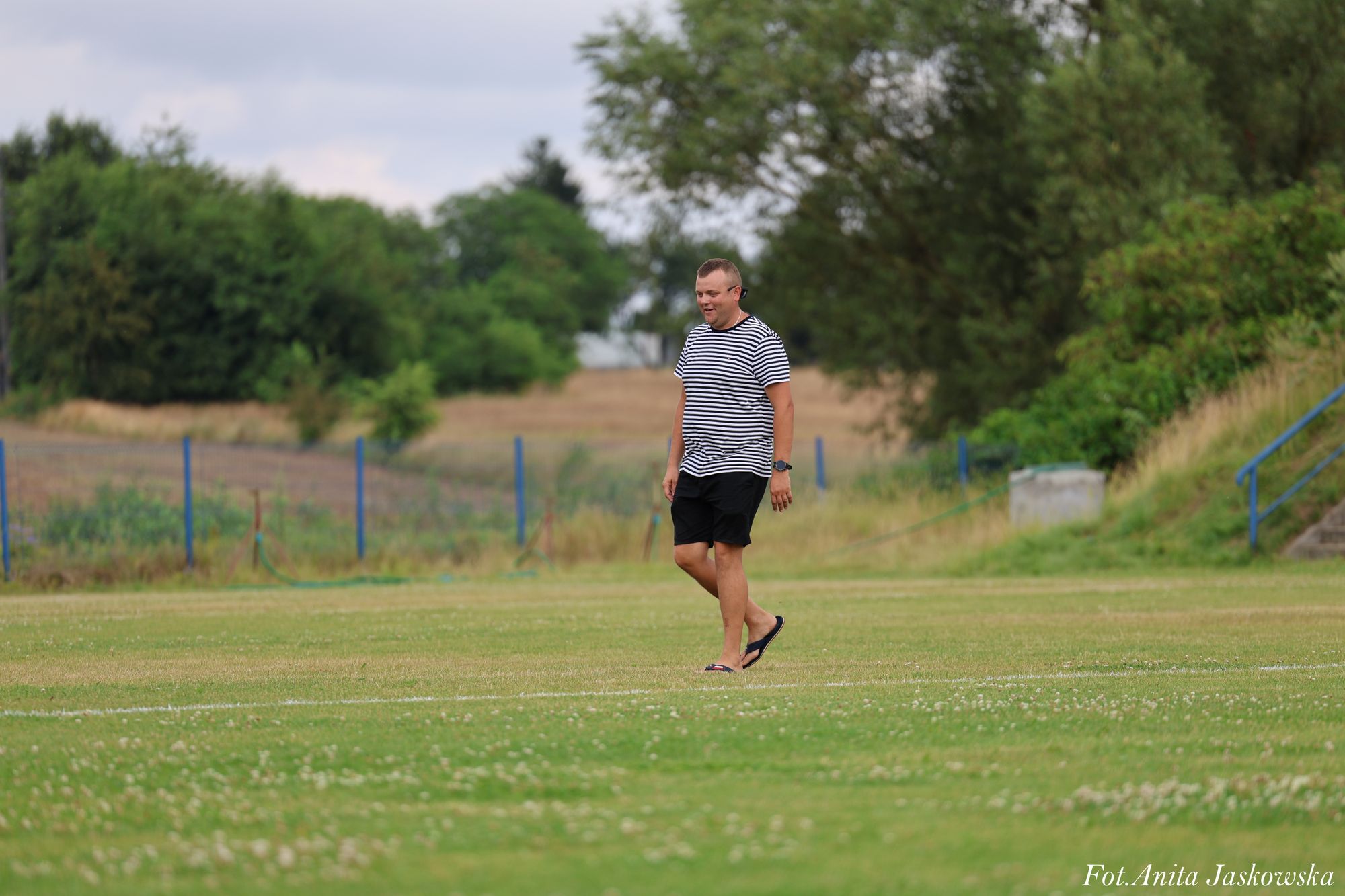Mężczyzna w czarno-białej pasiastej koszulce i czarnych spodenkach, spacerujący boso po zielonym boisku, w tle drzewa i ogrodzenie z niebieskimi słupkami.