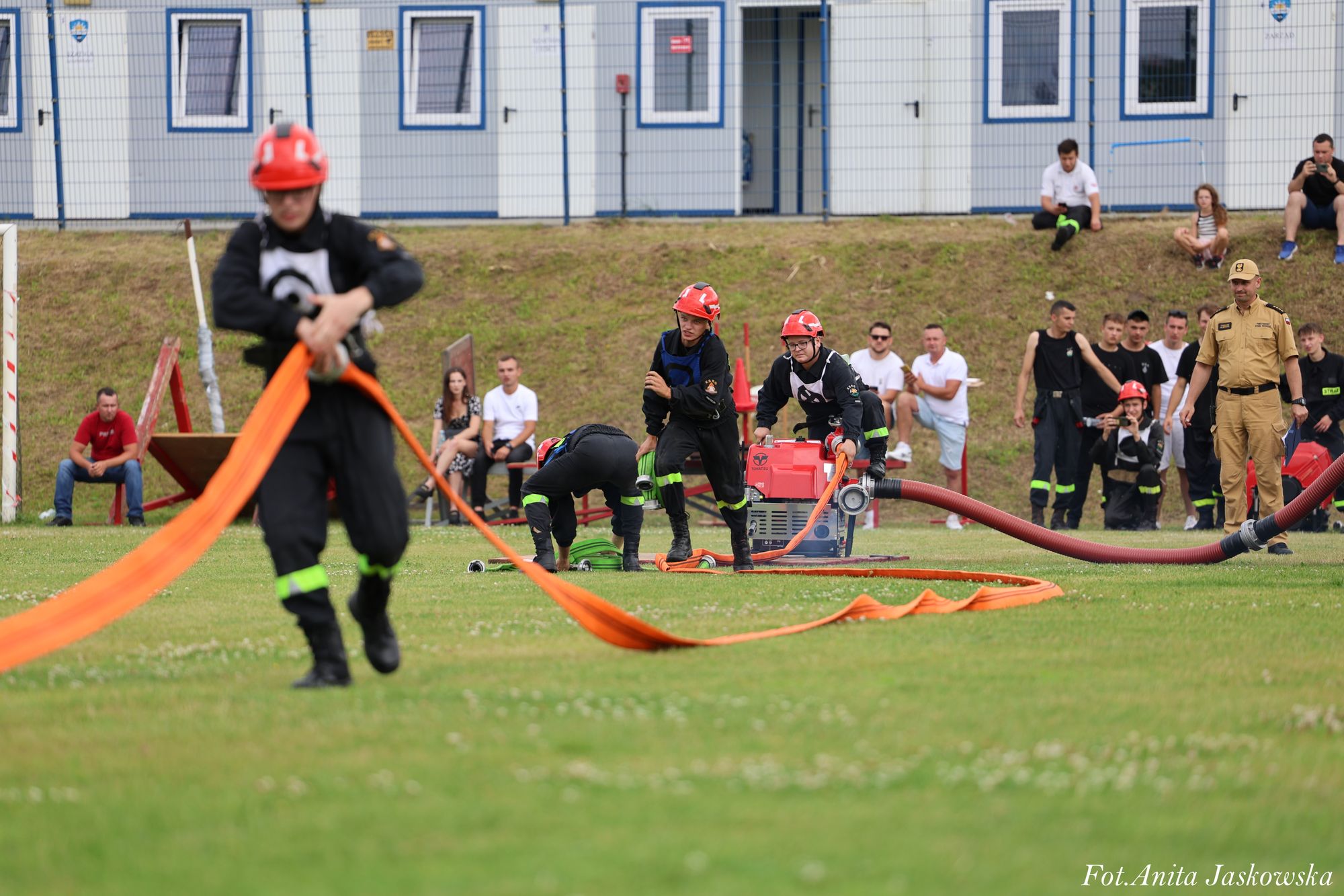 Młodzi strażacy w czarnych uniformach i czerwonych hełmach rozwijają pomarańczowe węże na zielonym boisku, w tle siedzą i stoją widzowie oraz budynki z oknami.