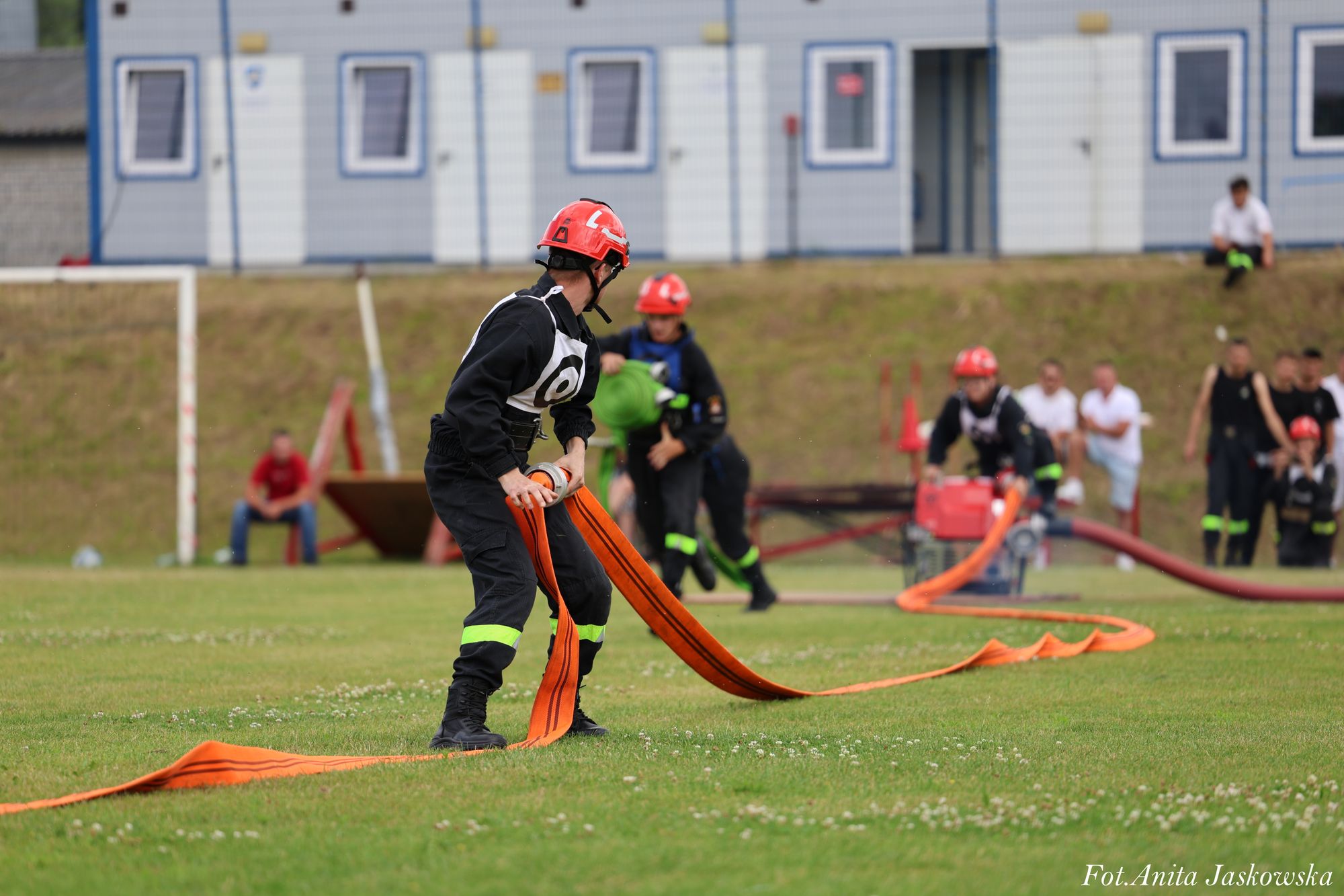 Mężczyzna w czarnym ubraniu z odblaskowymi paskami i czerwonym kasku rozwija pomarańczowy wąż na zielonej trawie, w tle inni ludzie i budynki.
