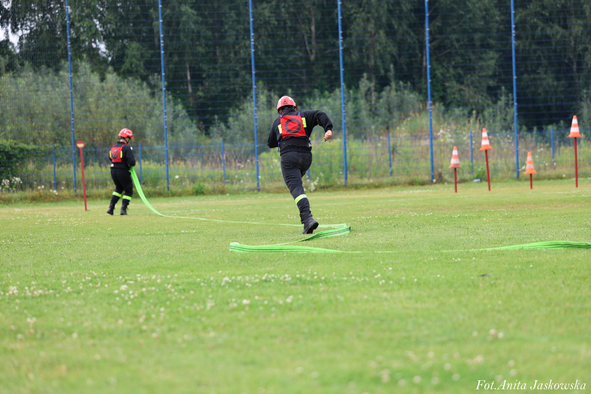 Dwóch strażaków w czarnych mundurach i czerwonych hełmach na zielonym boisku z rozłożonymi zielonymi wężami strażackimi i pachołkami po prawej stronie.