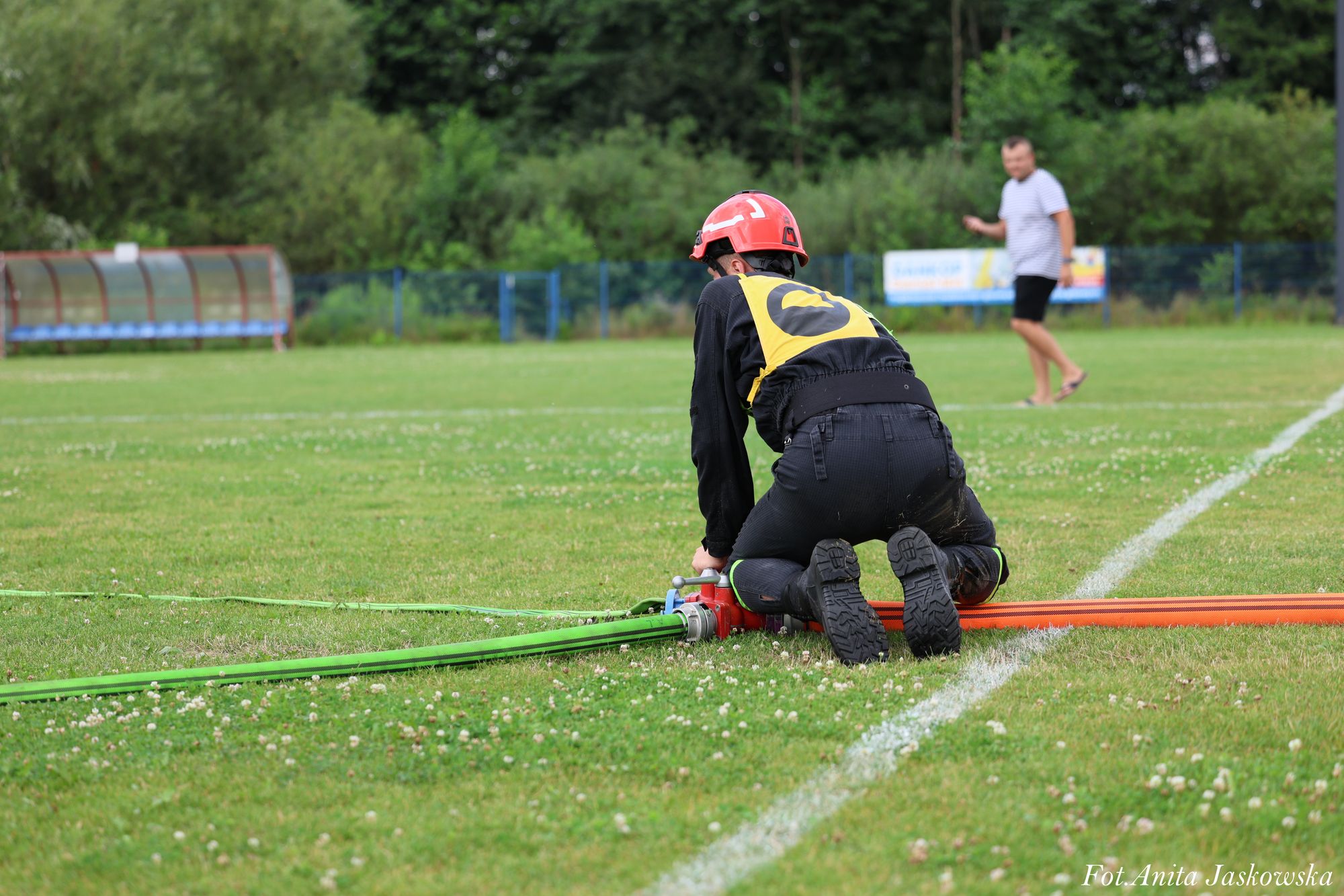 Osoba w czarnym ubraniu, czerwonym kasku i żółtej kamizelce klęczy na trawie, obsługując zielony i pomarańczowy wąż pożarniczy na boisku sportowym.