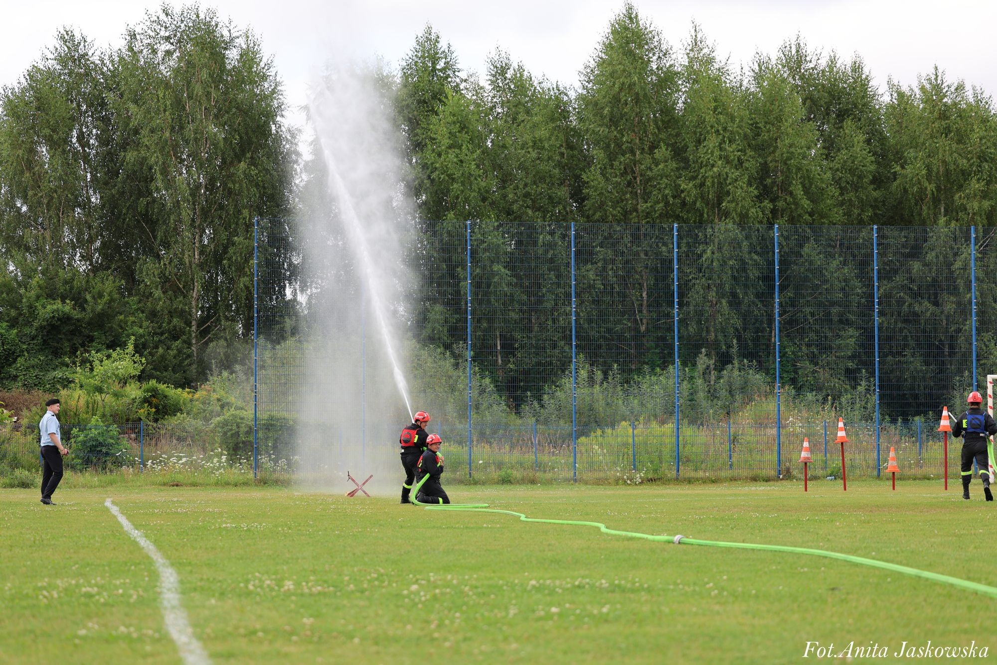 Strażacy w czarnych mundurach i czerwonych hełmach obsługują zielony wąż strażacki na zielonym boisku, woda tryska pionowo, w tle drzewa i ogrodzenie z siatki.