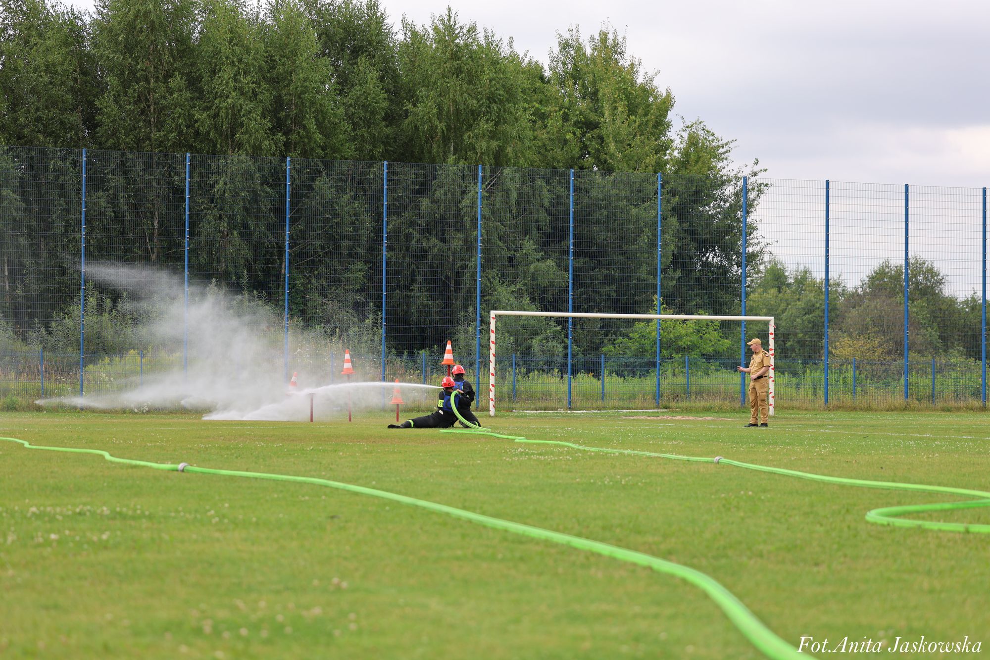 Dwoje strażaków w czarnych mundurach i czerwonych hełmach używa zielonego węża strażackiego na zielonym boisku, mężczyzna w beżowym mundurze stoi obok bramki, w tle ogrodzenie i drzewa.