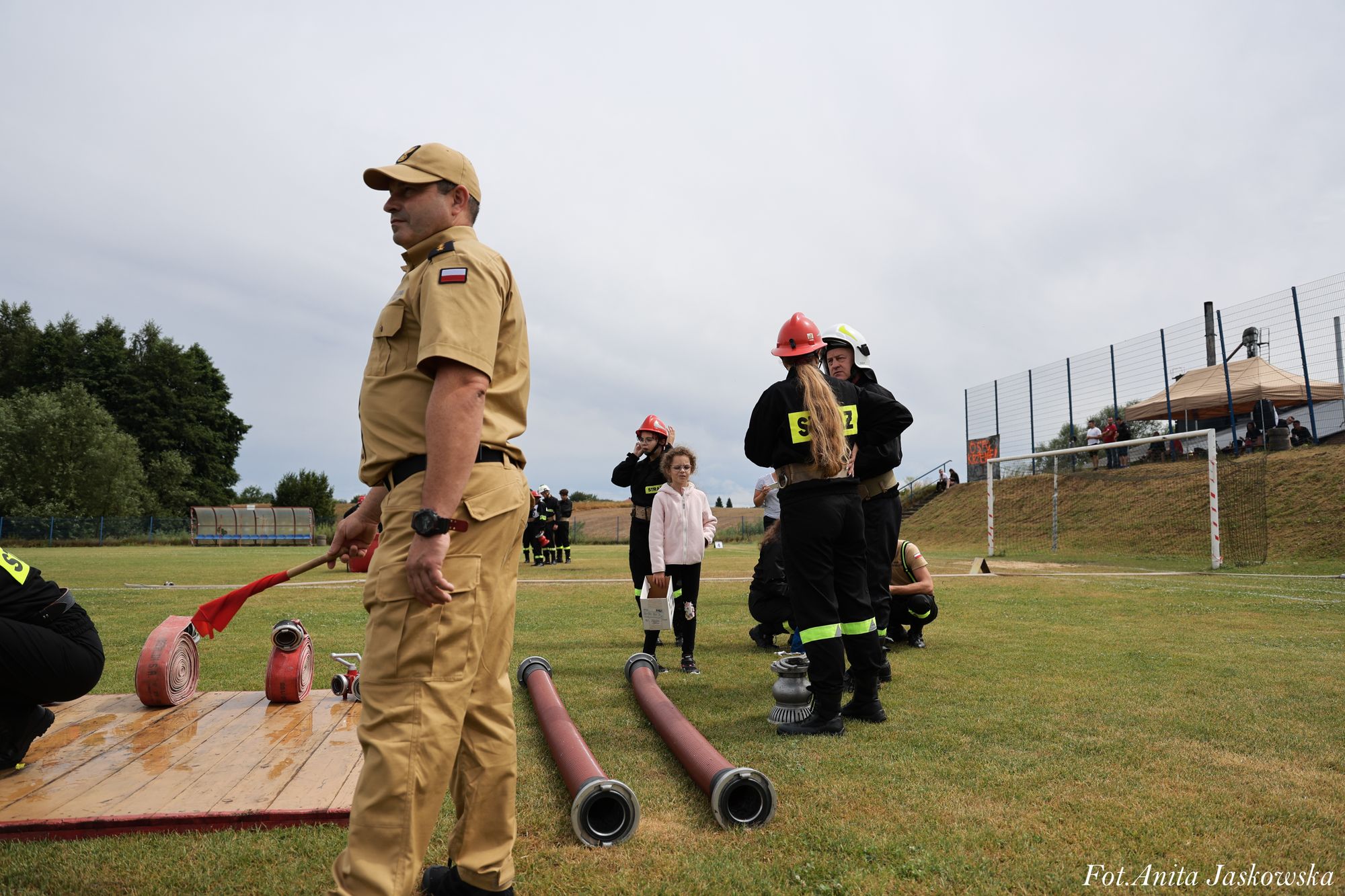 Mężczyzna w piaskowym mundurze z polską flagą na rękawie trzymający czerwoną flagę, osoby w czarnych mundurach strażackich i czerwonych hełmach na trawie z wężami strażackimi.