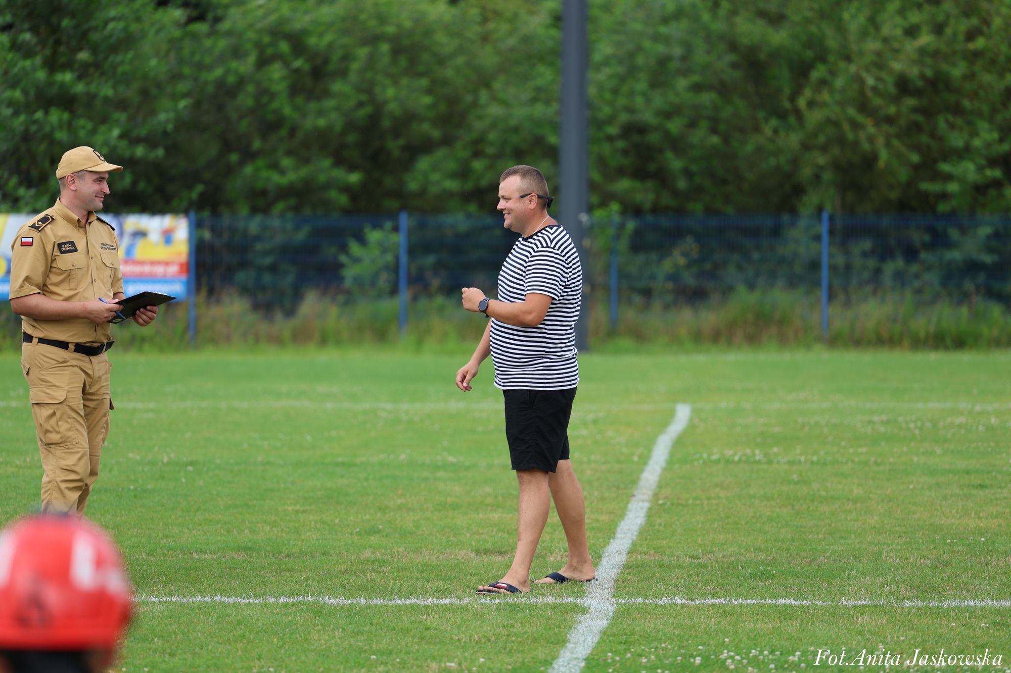 Dwóch mężczyzn na zielonej trawie boiska, jeden w beżowym mundurze z notatnikiem, drugi w czarno-białej koszulce w paski i czarnych szortach.