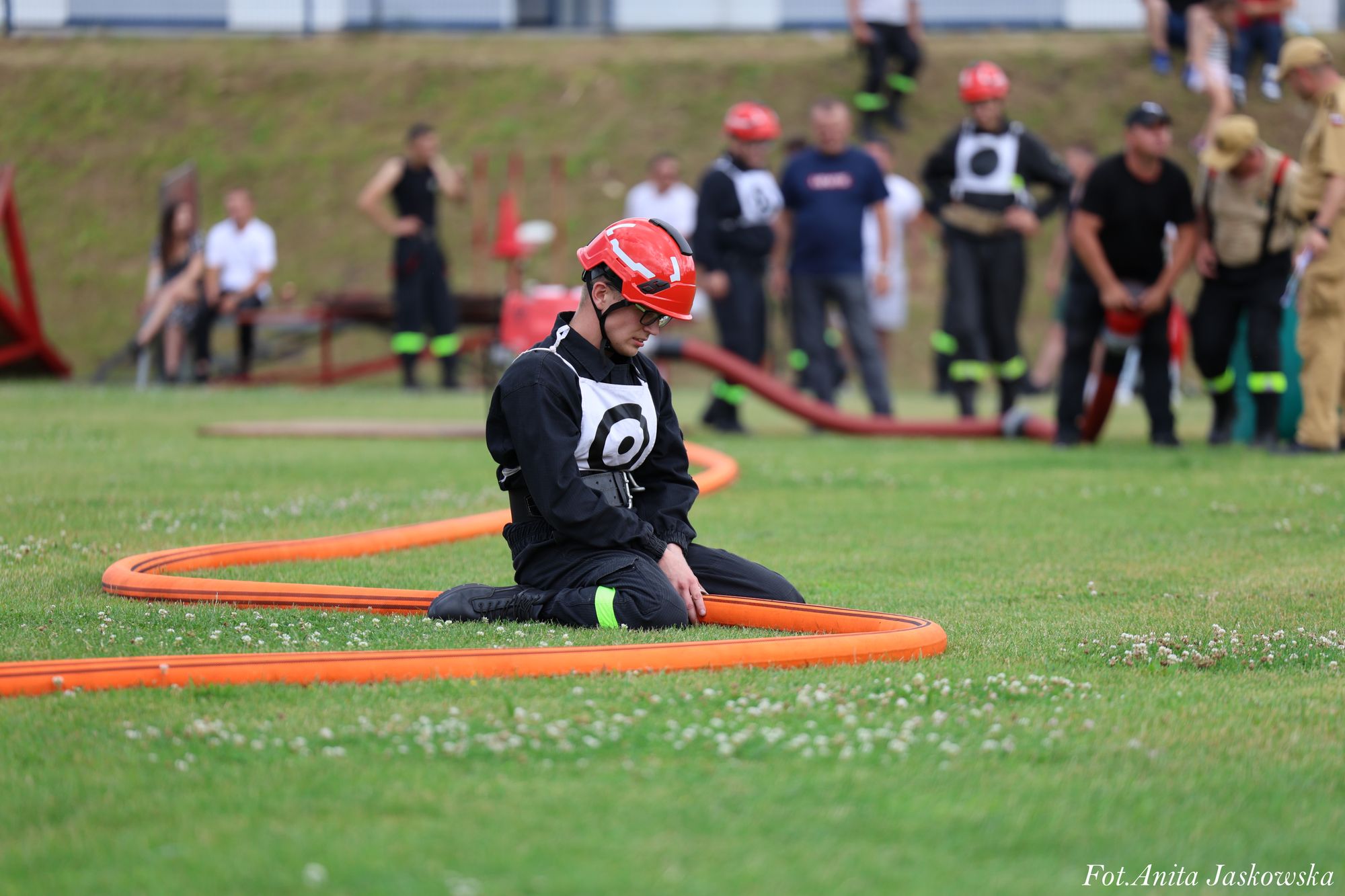 Mężczyzna w czarnym ubraniu i czerwonym kasku klęczy na zielonej trawie, trzyma pomarańczowy wąż strażacki, w tle rozmyta grupa ludzi.