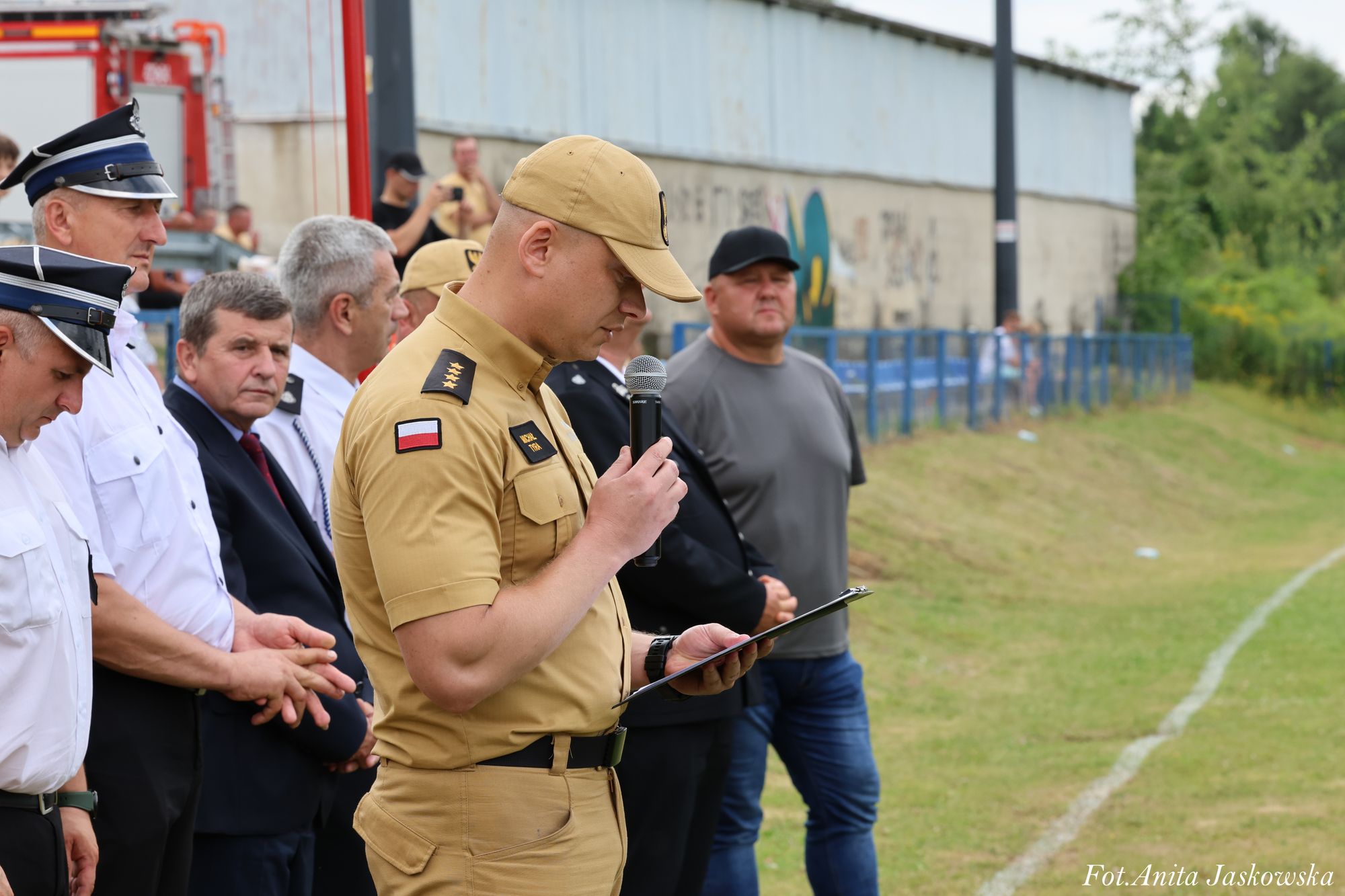 Mężczyzna w beżowym mundurze i czapce trzyma mikrofon i czyta z teczki, obok mężczyźni w białych koszulach i granatowych garniturach, na zewnątrz trawiaste boisko.