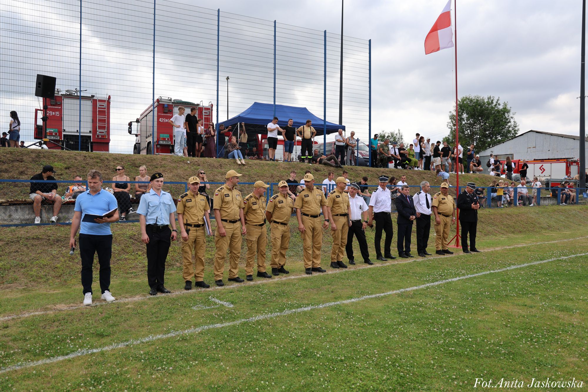 Grupa mężczyzn w mundurach strażackich i innych strojach stoi w rzędzie na trawie, za nimi widownia i dwa czerwone wozy strażackie, flaga czerwona z białym kwadratem.