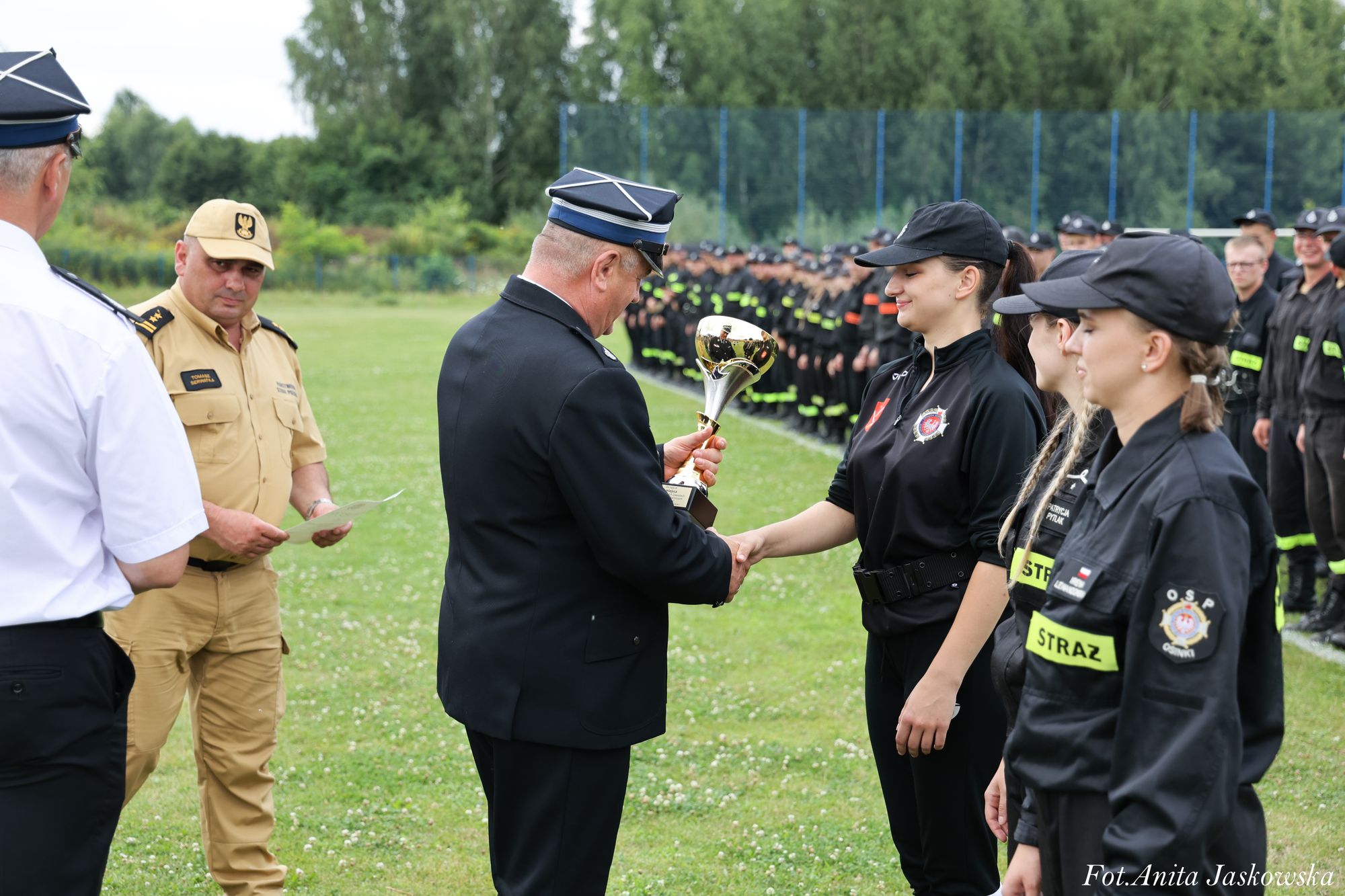 Mężczyzna w granatowym mundurze wręcza puchar kobiecie w czarnym ubraniu z napisem "STRAŻ", obok stoją inne kobiety w podobnym mundurze, w tle szereg osób w mundurach strażackich na zielonym polu.