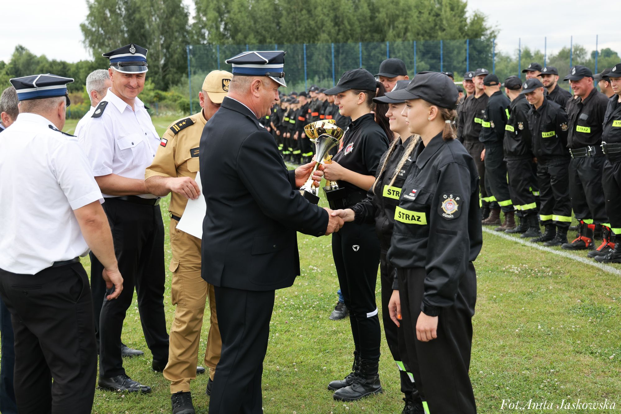 Grupa umundurowanych strażaków na zielonym boisku, mężczyzna w czarnym mundurze wręcza puchar młodej kobiecie w czarnym stroju z napisem STRAŻ.