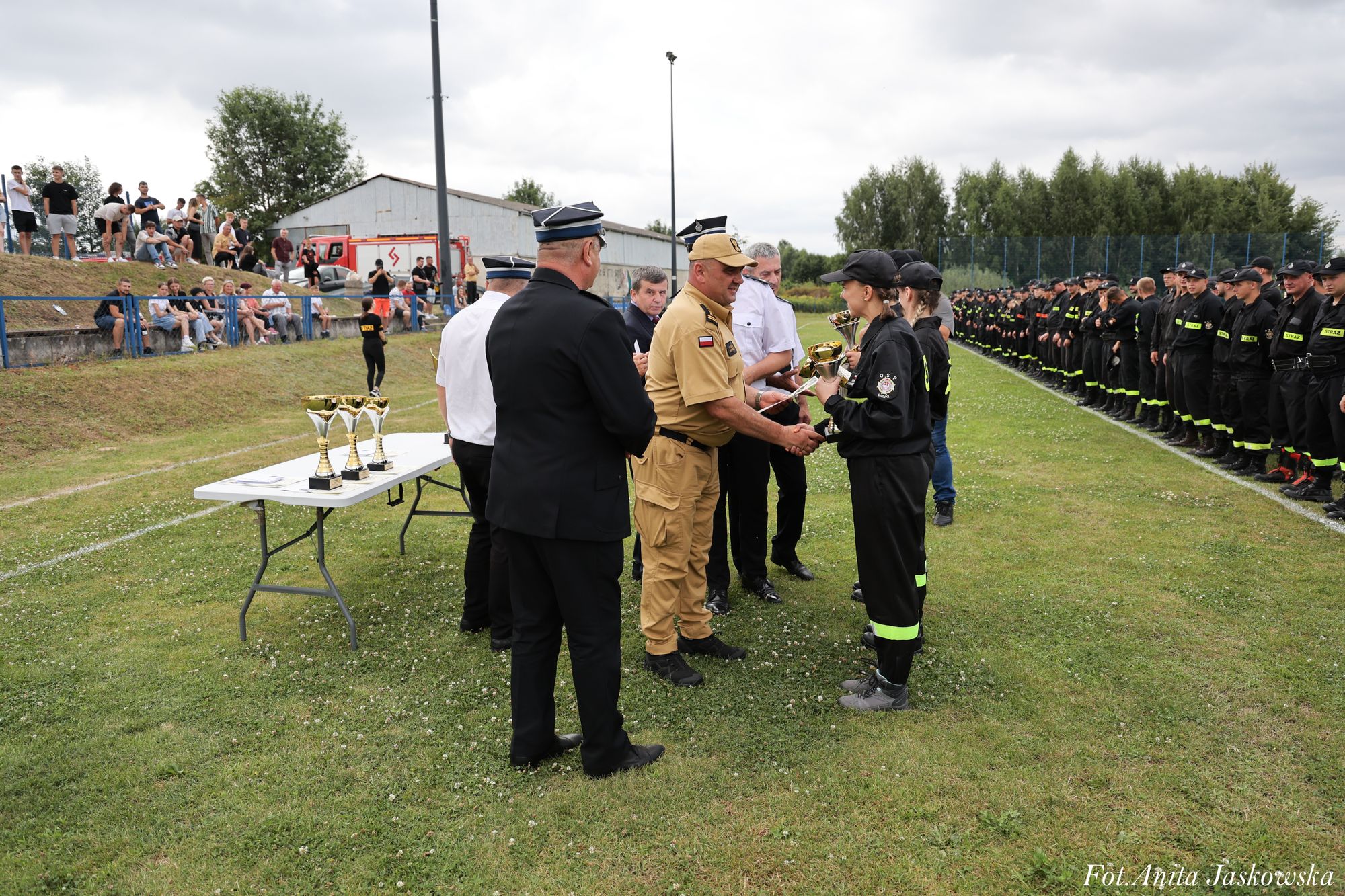 Osoby w mundurach na trawie, kobiety w czarnych uniformach z odblaskowymi pasami odbierają puchary, w tle rząd stojących osób w czarnych mundurach i widzowie na trybunach.