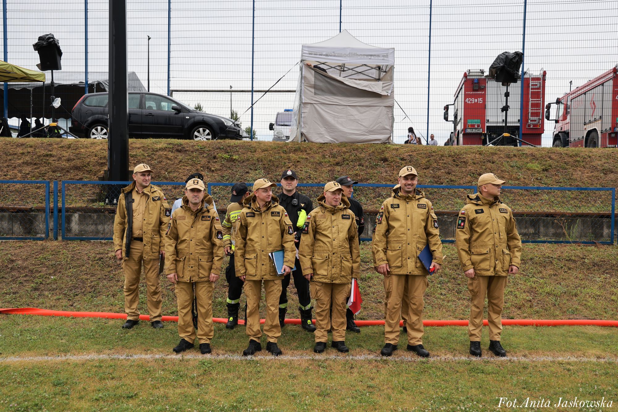 Siedmiu mężczyzn ubranych w beżowe mundury z czarnymi butami stoi na trawie, za nimi niebieska barierka, samochody, namiot i czerwone wozy strażackie.