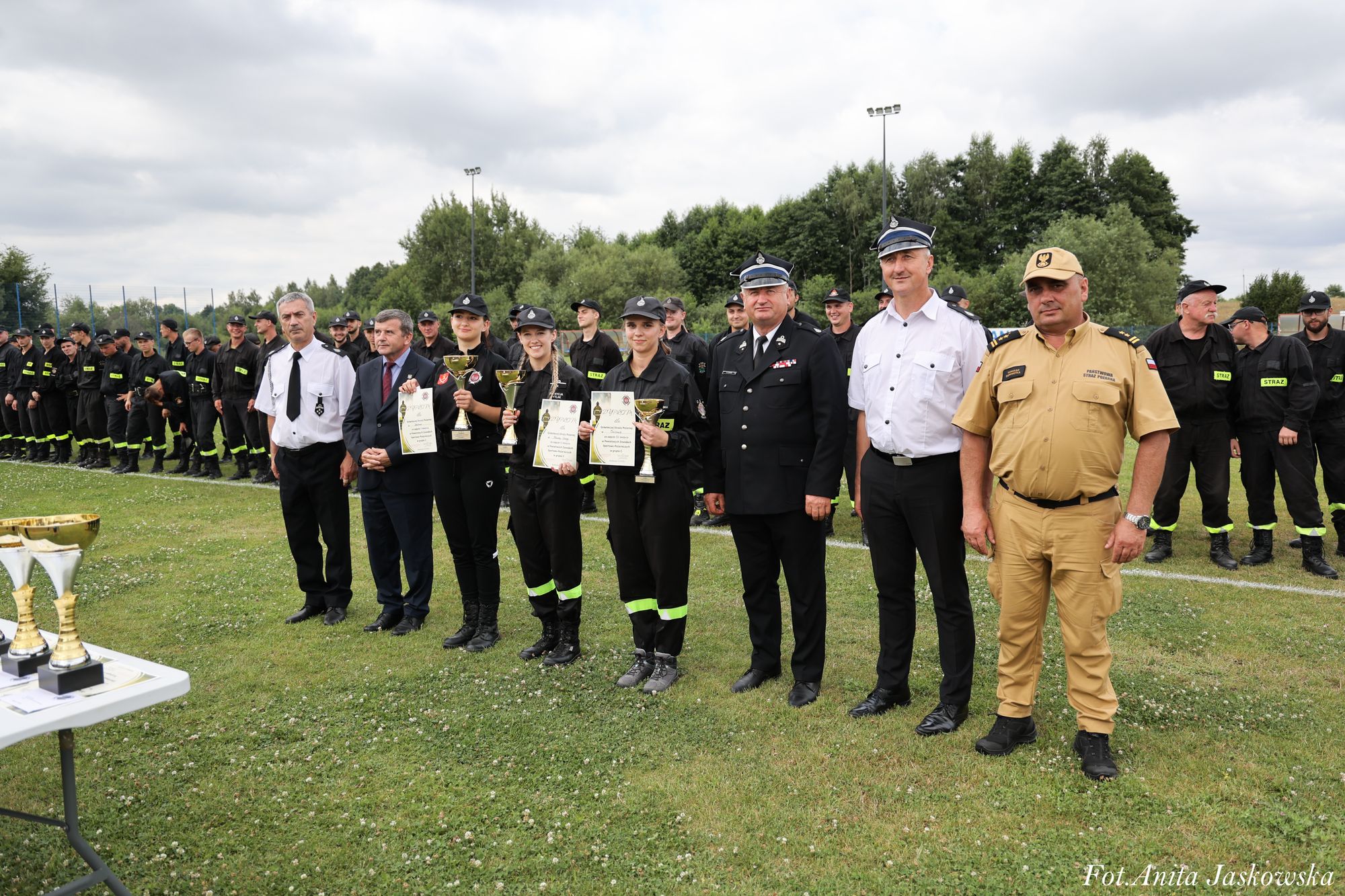 Grupa osób w mundurach strażackich i oficjalnych stoi na trawie, trzy kobiety trzymają puchary i dyplomy, w tle drzewa i pochmurne niebo.