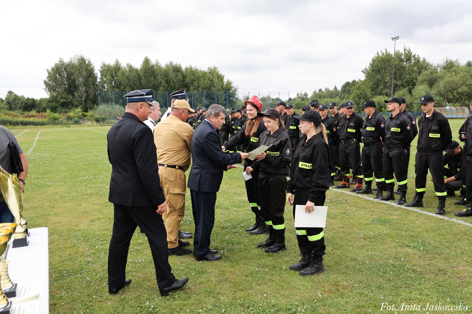 Grupa osób w mundurach strażackich stoi na trawie, trzy kobiety odbierają dyplomy od mężczyzn w garniturach i mundurach, w tle drzewa i ogrodzenie boiska.