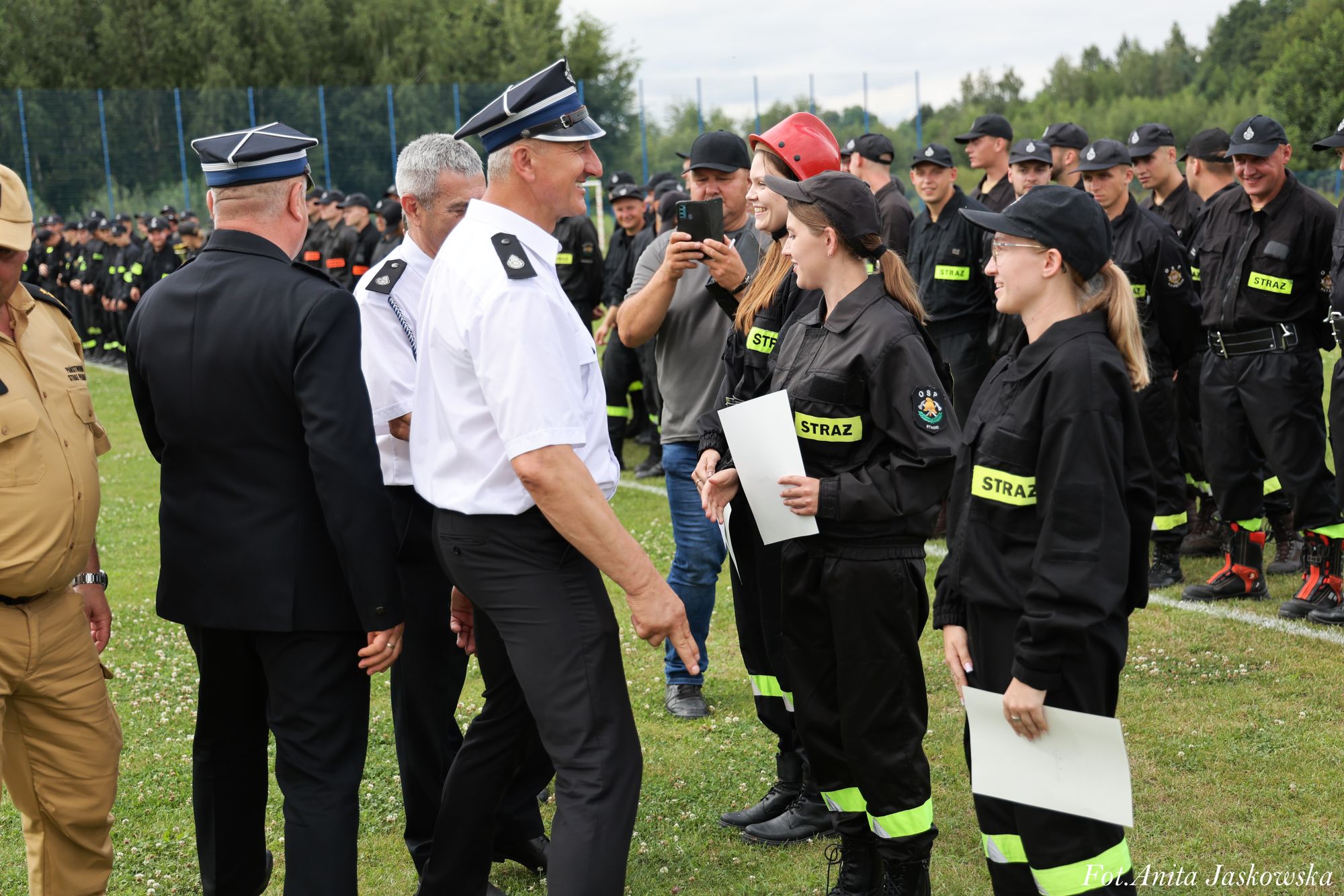 Mężczyźni w mundurach wręczają dokumenty młodym kobietom w czarnych strojach z napisem "STRAŻ", jedna kobieta w czerwonym kasku, w tle grupa osób w podobnych strojach na trawie.