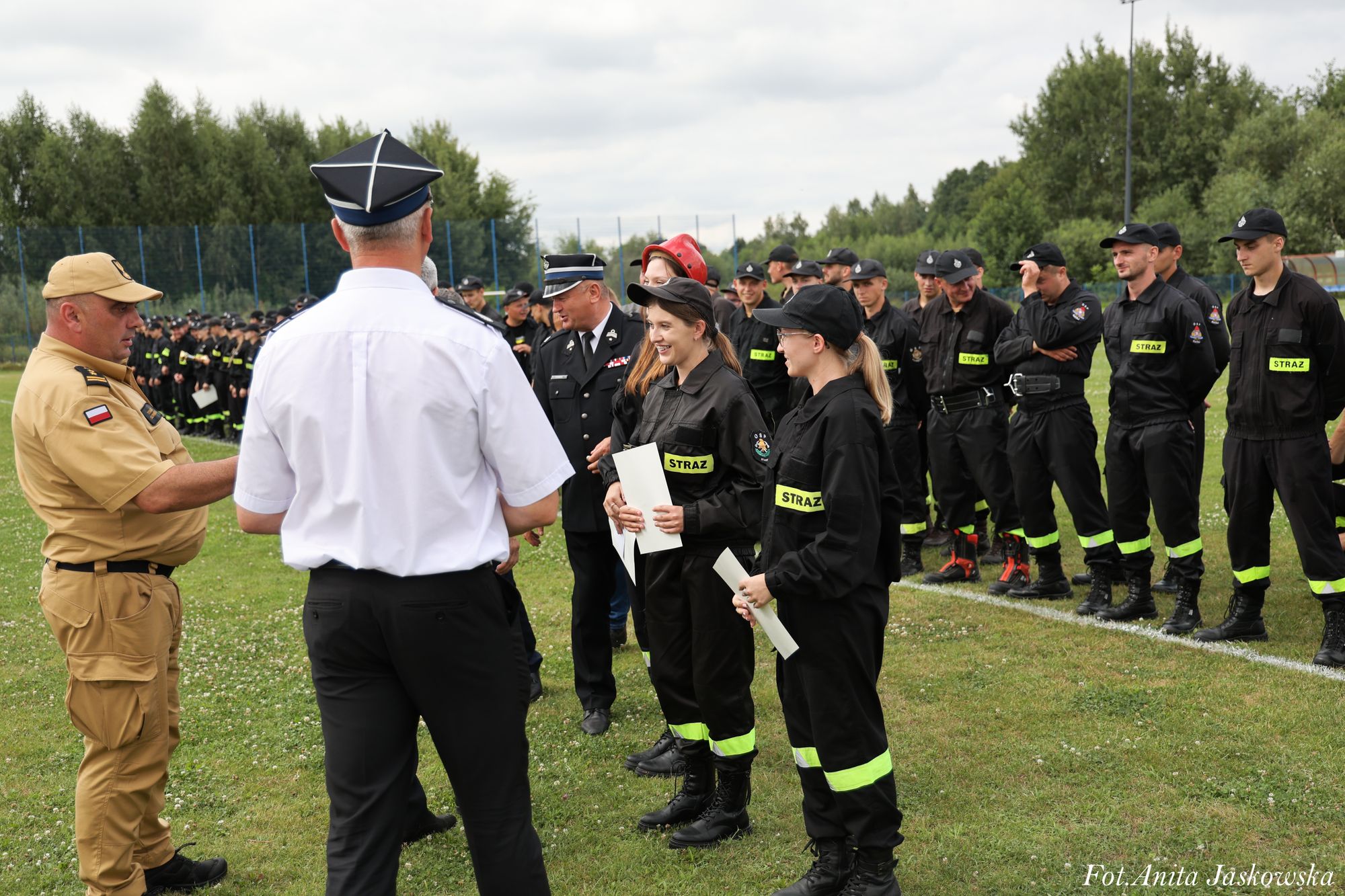 Grupa strażaków w czarnych mundurach z żółtymi odblaskami stoi na trawie, dwie kobiety z dokumentami rozmawiają z mężczyznami w mundurach, w tle drzewa i ogrodzenie.