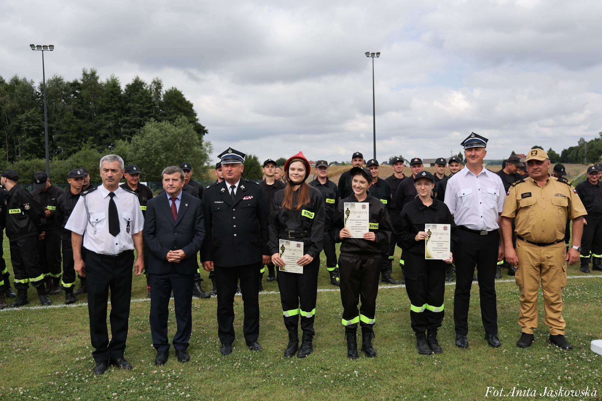 Grupa osób na trawie, mężczyźni w mundurach, kobiety w czarnych strojach strażackich z odblaskowymi wstawkami, trzy kobiety trzymają dyplomy.