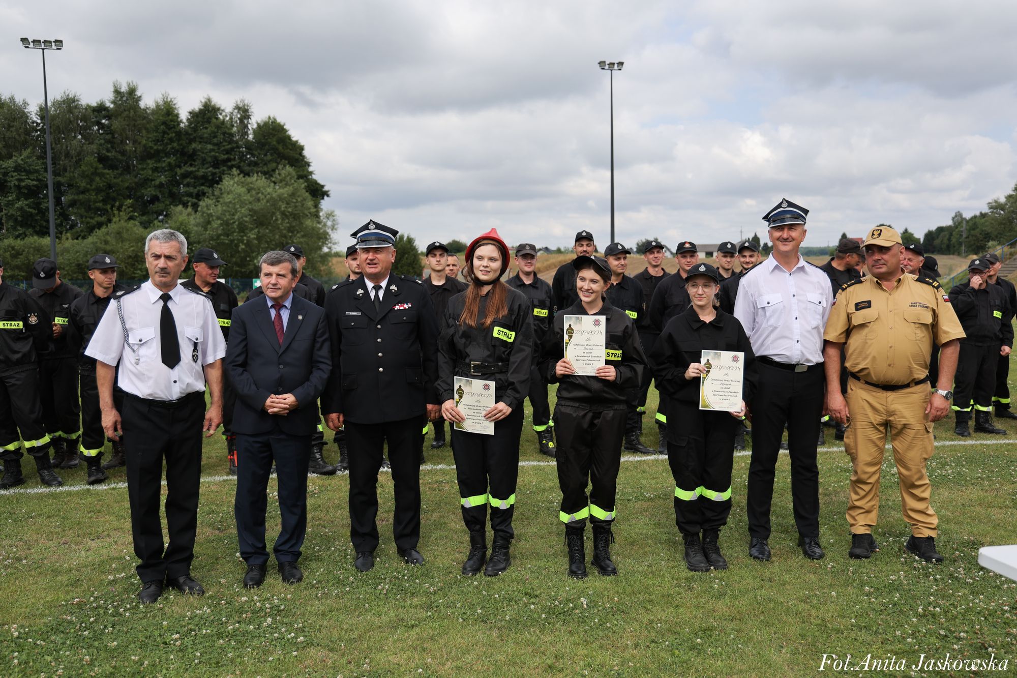 Grupa osób w mundurach strażackich i oficjalnych na trawie, trzy kobiety w czarnych kombinezonach z żółtymi odblaskami trzymają dyplomy, za nimi inni strażacy.