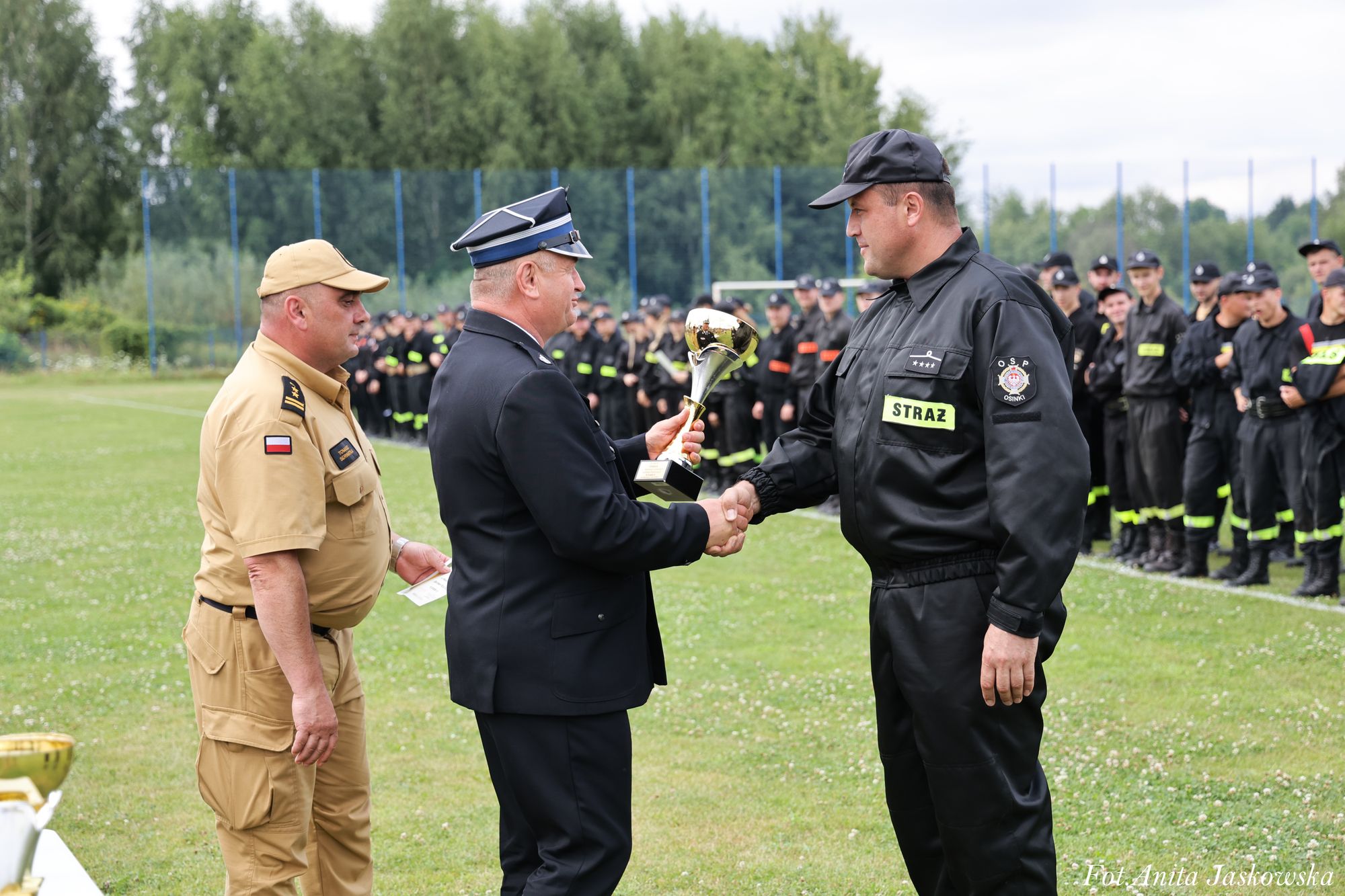Mężczyzna w czarnym mundurze wręcza puchar mężczyźnie w czarnym mundurze strażackim z napisem STRAŻ, obok stoi mężczyzna w beżowym mundurze, w tle grupa osób w czarnych mundurach na trawie.