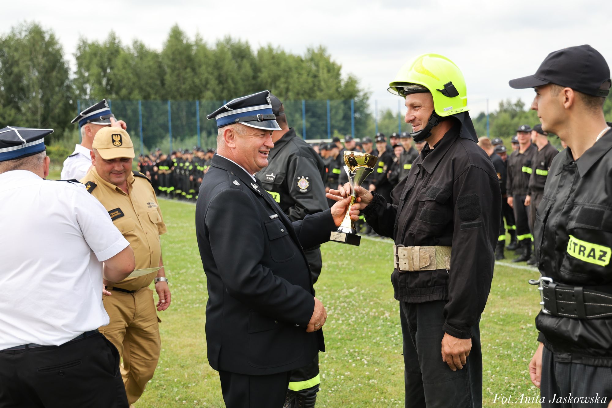 Mężczyzna w mundurze wręcza puchar strażakowi w czarnym stroju i żółtym hełmie, na tle grupy osób w czarnych mundurach strażackich na zielonej trawie.