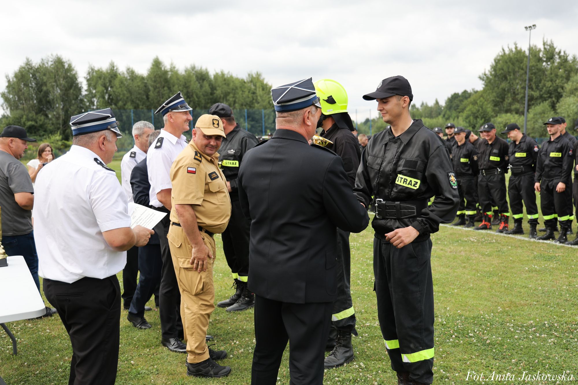 Grupa ludzi na trawie, mężczyźni w mundurach strażackich czarnych i białych, jeden w beżowym mundurze, kilka osób w czapkach, w tle drzewa i słup oświetleniowy.