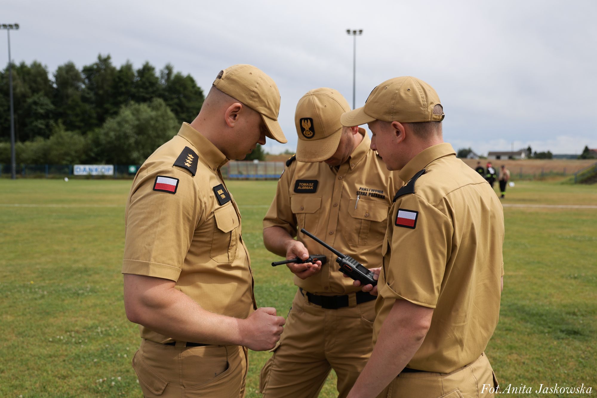 Trzech mężczyzn w beżowych mundurach z naszywkami polskiej flagi i czapkach, stojących na zielonym boisku, trzymających krótkofalówki.