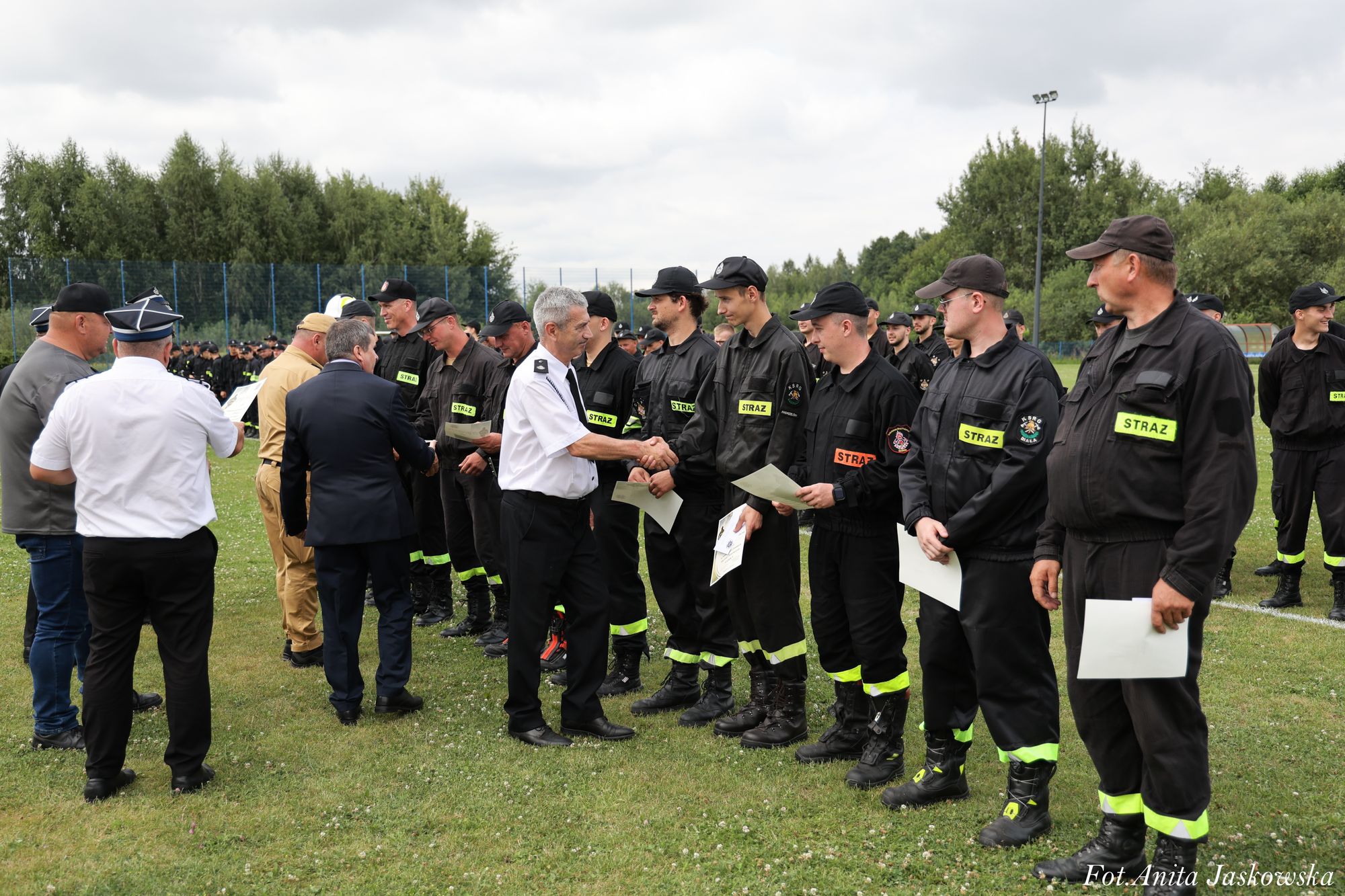 Grupa mężczyzn w czarnych mundurach z napisem "STRAŻ" stoi na zielonej trawie, kilku otrzymuje dokumenty i podaje ręce mężczyznom w białych i granatowych koszulach, w tle drzewa i ogrodzenie.