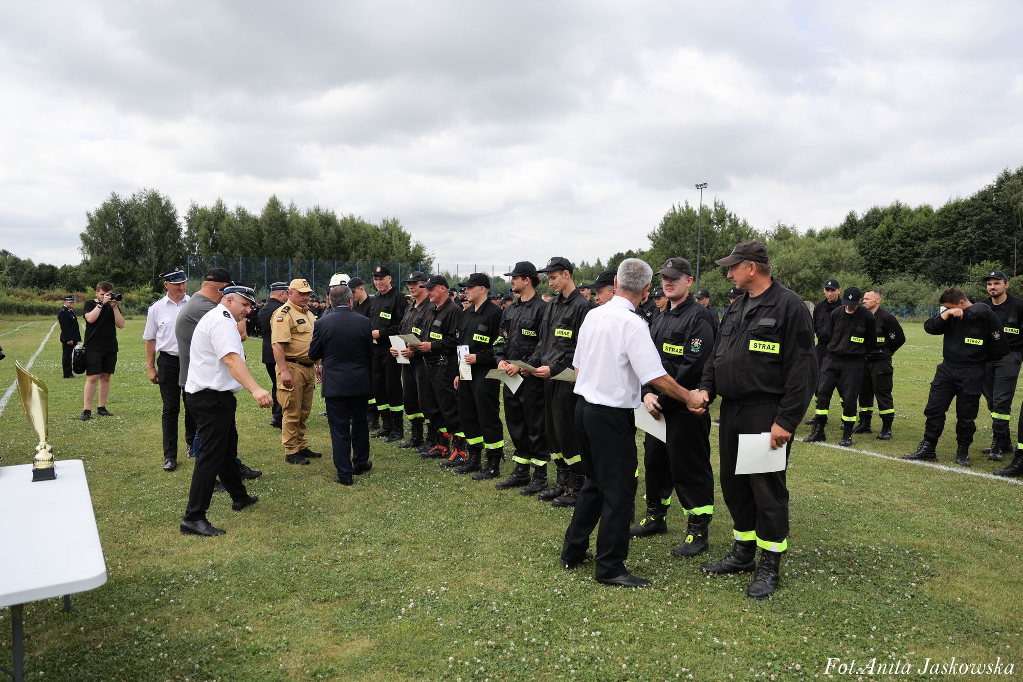 Grupa mężczyzn w mundurach strażackich i oficjalnych na zielonym boisku, niektórzy trzymają dyplomy, na stole trofeum, zachmurzone niebo.