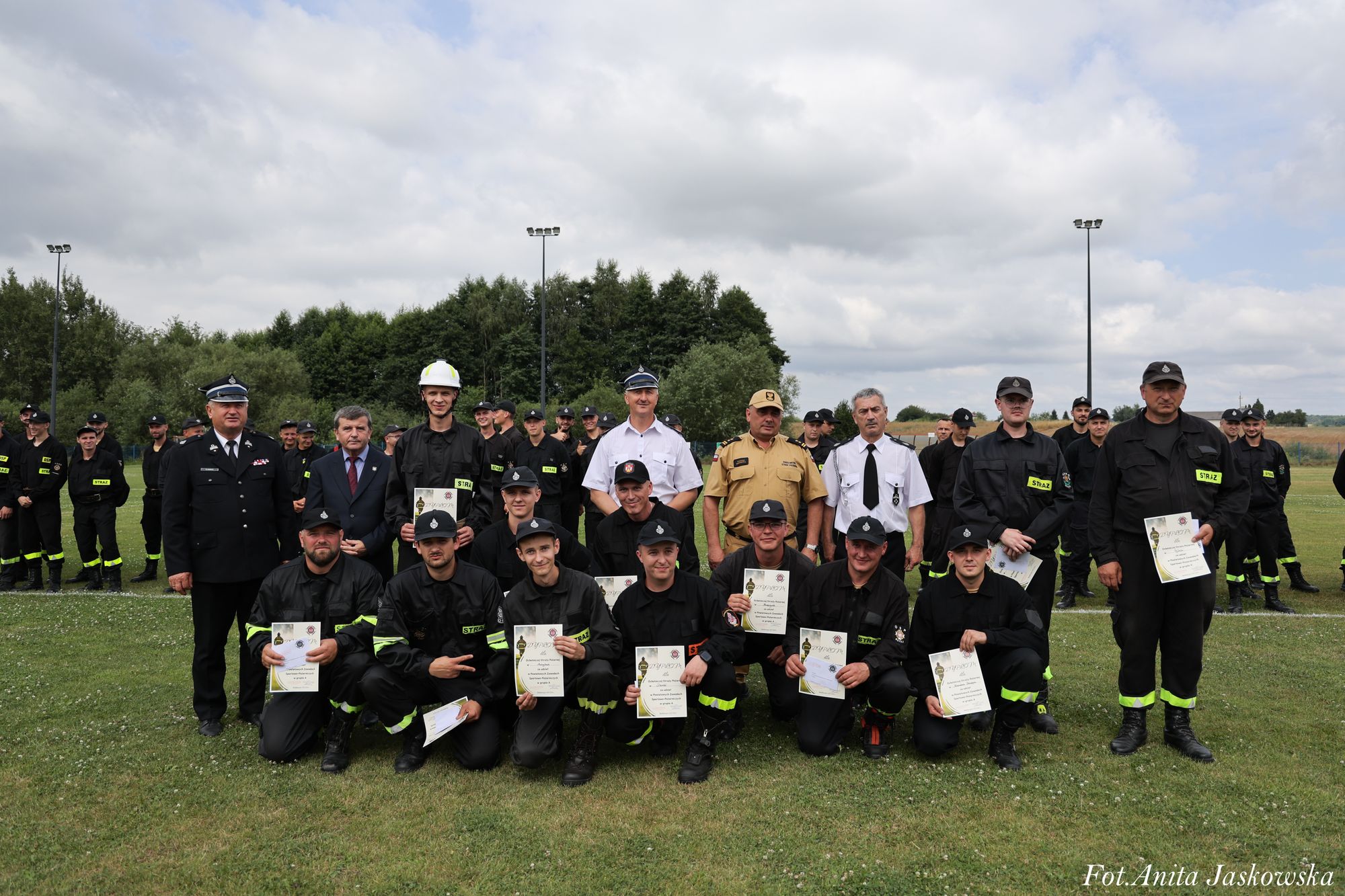 Grupa mężczyzn w mundurach strażackich i oficerskich, niektórzy trzymają dyplomy, stoją i klęczą na trawie, w tle drzewa i pochmurne niebo.