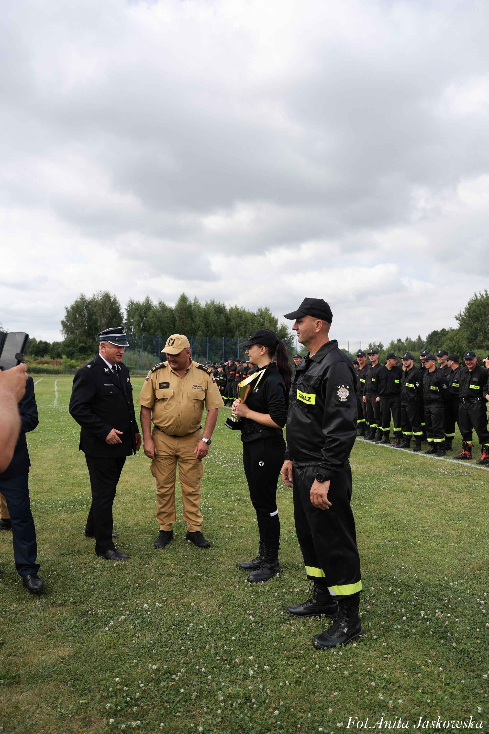 Grupa osób w mundurach strażackich i ratowniczych na trawiastej łące, jedna kobieta trzyma złoty puchar, w tle linia osób w czarnych mundurach ze żółtymi napisami.