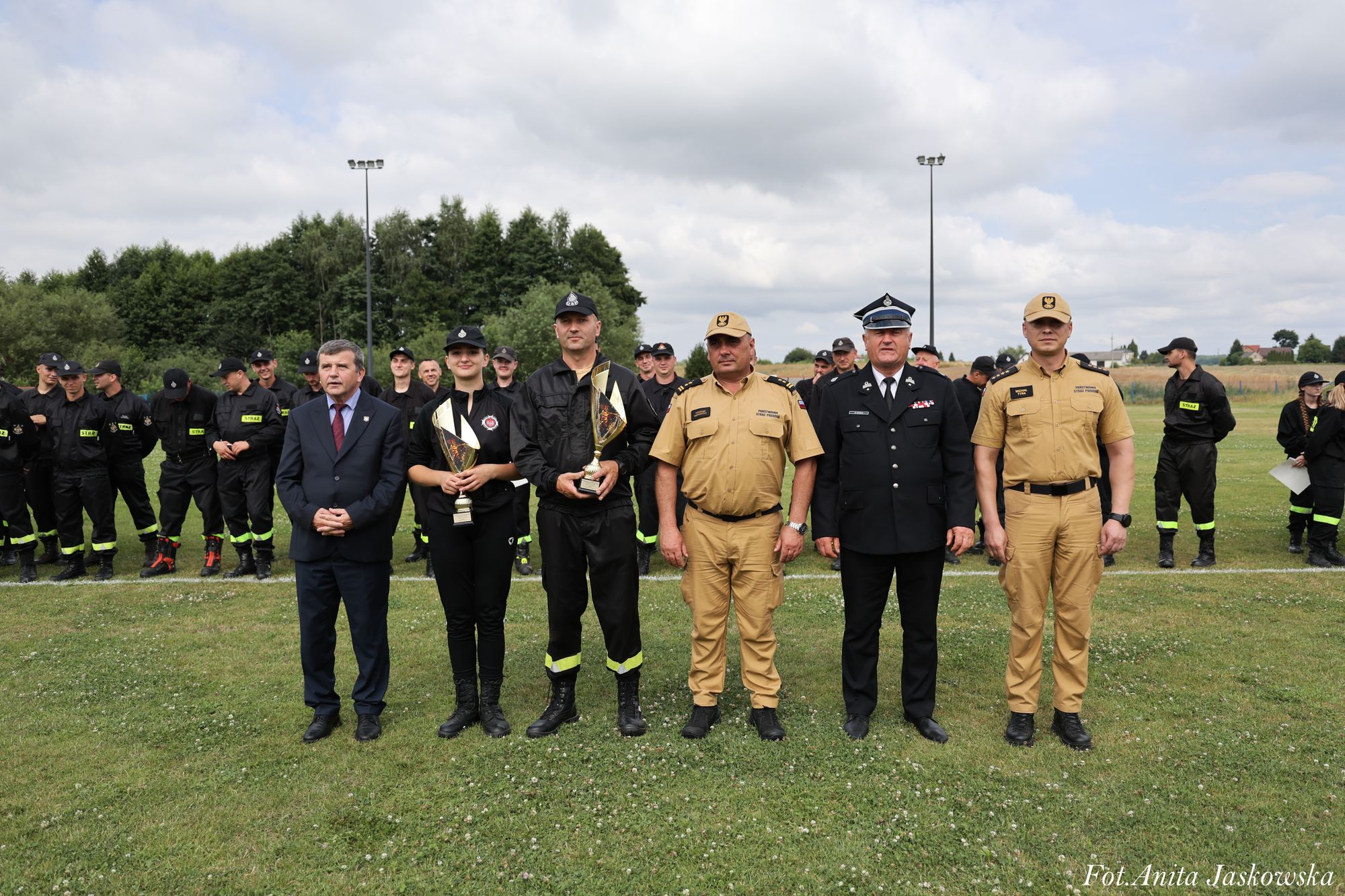 Grupa osób w mundurach strażackich i cywilnych stojąca na zielonym boisku, dwie osoby trzymają złote puchary, w tle drzewa i pochmurne niebo.
